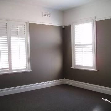 Empty room with brown walls, white trim, two shuttered windows, and dark carpet. — S D Maintenance In Point Clare, NSW