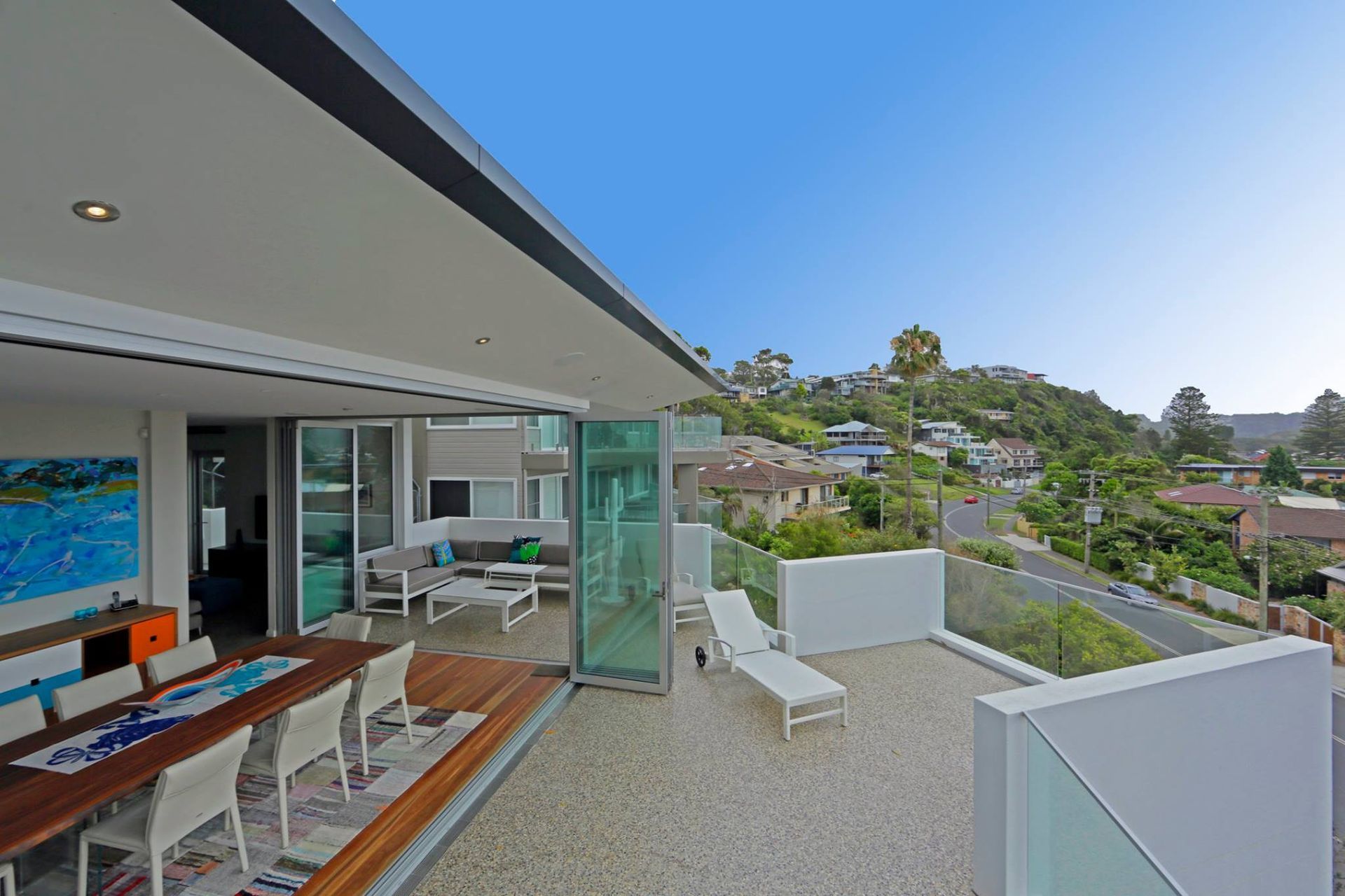 Modern home with open-air deck overlooking a neighborhood and the sky. Outdoor dining table, lounge chairs, and partially open glass doors. — S D Maintenance In Point Clare, NSW