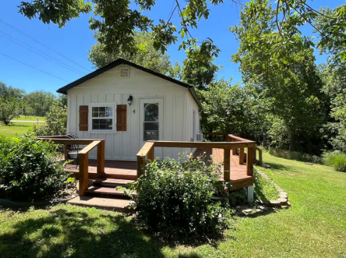 A small white house with a wooden deck in the middle of a grassy field.