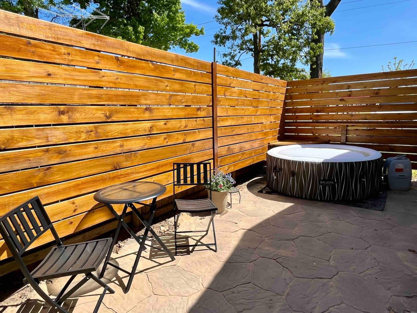 A wooden fence surrounds a patio with a hot tub and a table and chairs.
