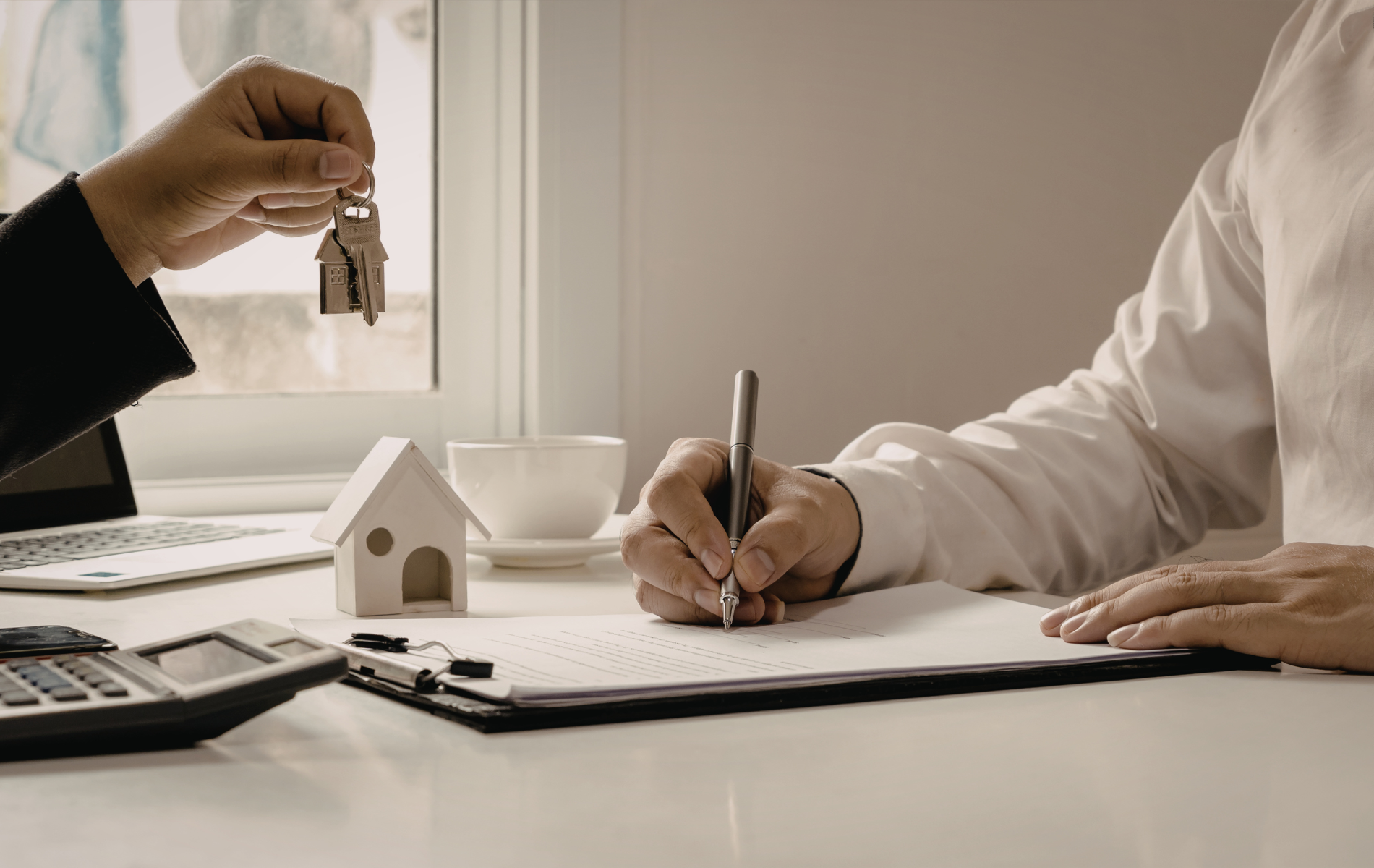 Person signing documents at a desk as another hands over house keys.