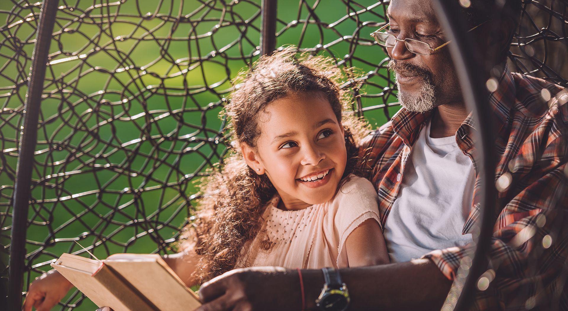 A man and a little girl are sitting in a wicker chair reading a book.