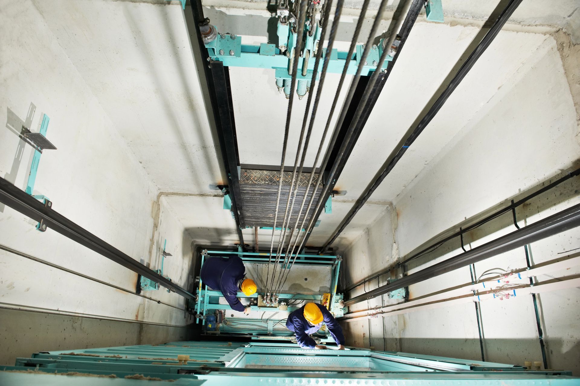Dos trabajadores en el hueco de una escalera, cerca de la plataforma de un ascensor, rodeados de cables y raíles.