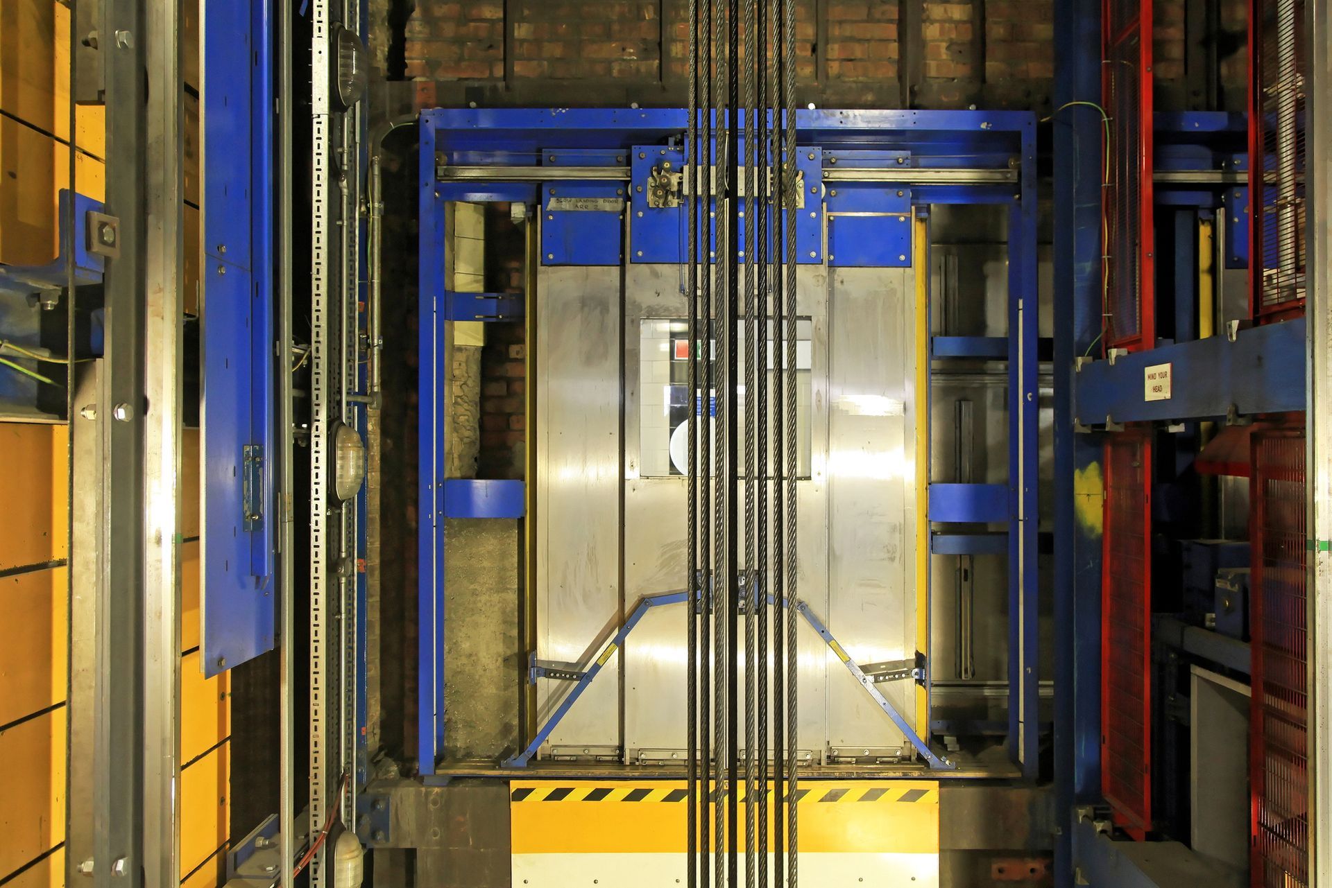 Hueco de ascensor industrial con estructura metálica azul y plataforma amarilla en un entorno similar a un almacén.