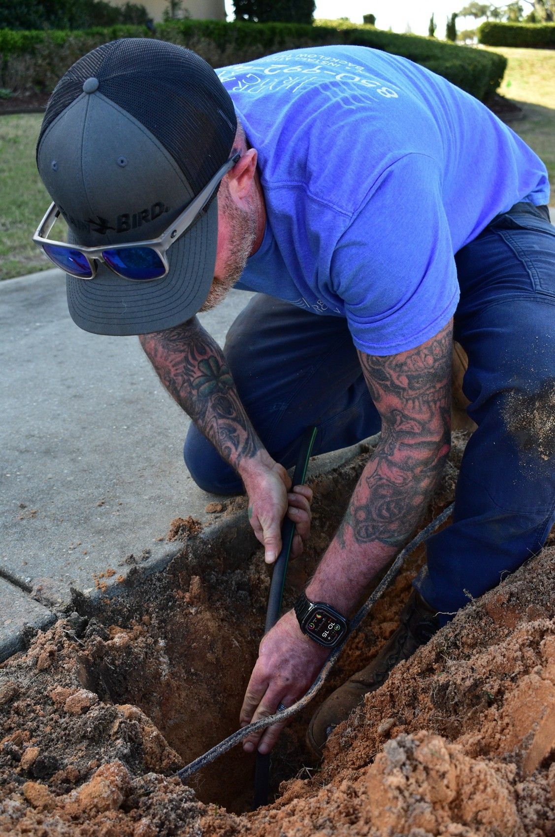 Man in blue shirt, kneeling, working in a dirt trench, possibly repairing something.
