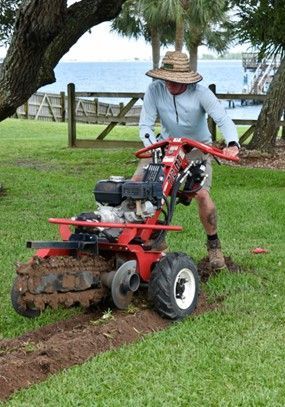 Man using a red ditch witch to dig a trench in a grassy yard near water.