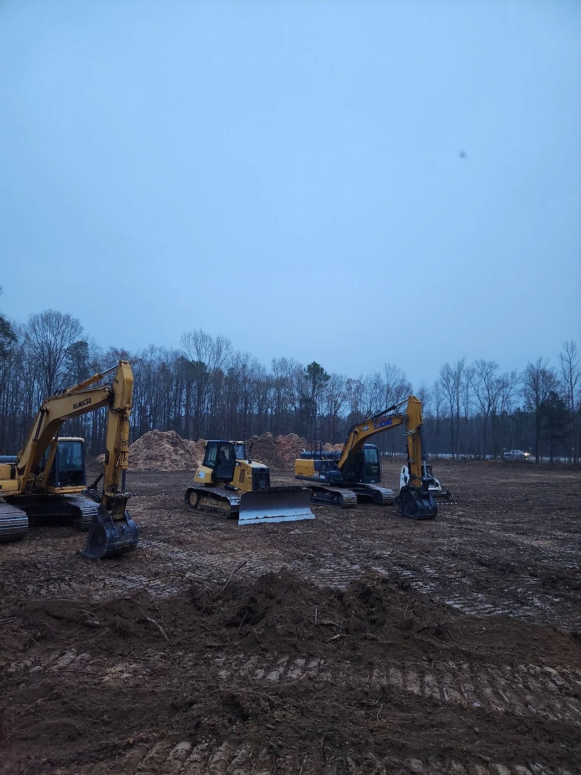 Three yellow construction excavators on a muddy field under a cloudy sky.