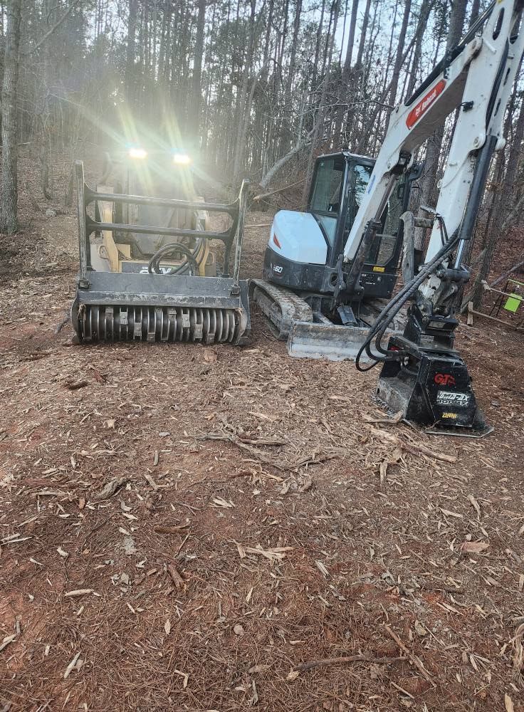 Bobcat excavator with brush cutter in a wooded area, ground covered in wood chips.