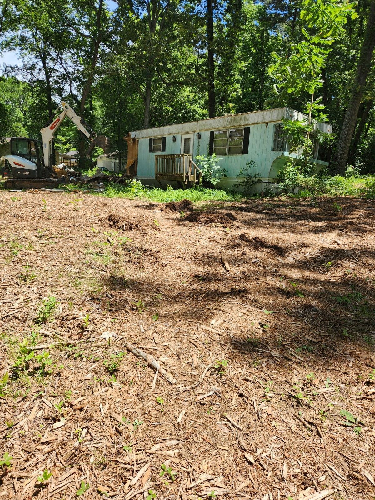 A mobile home being demolished by an excavator in a wooded area. Brown earth covers the foreground.