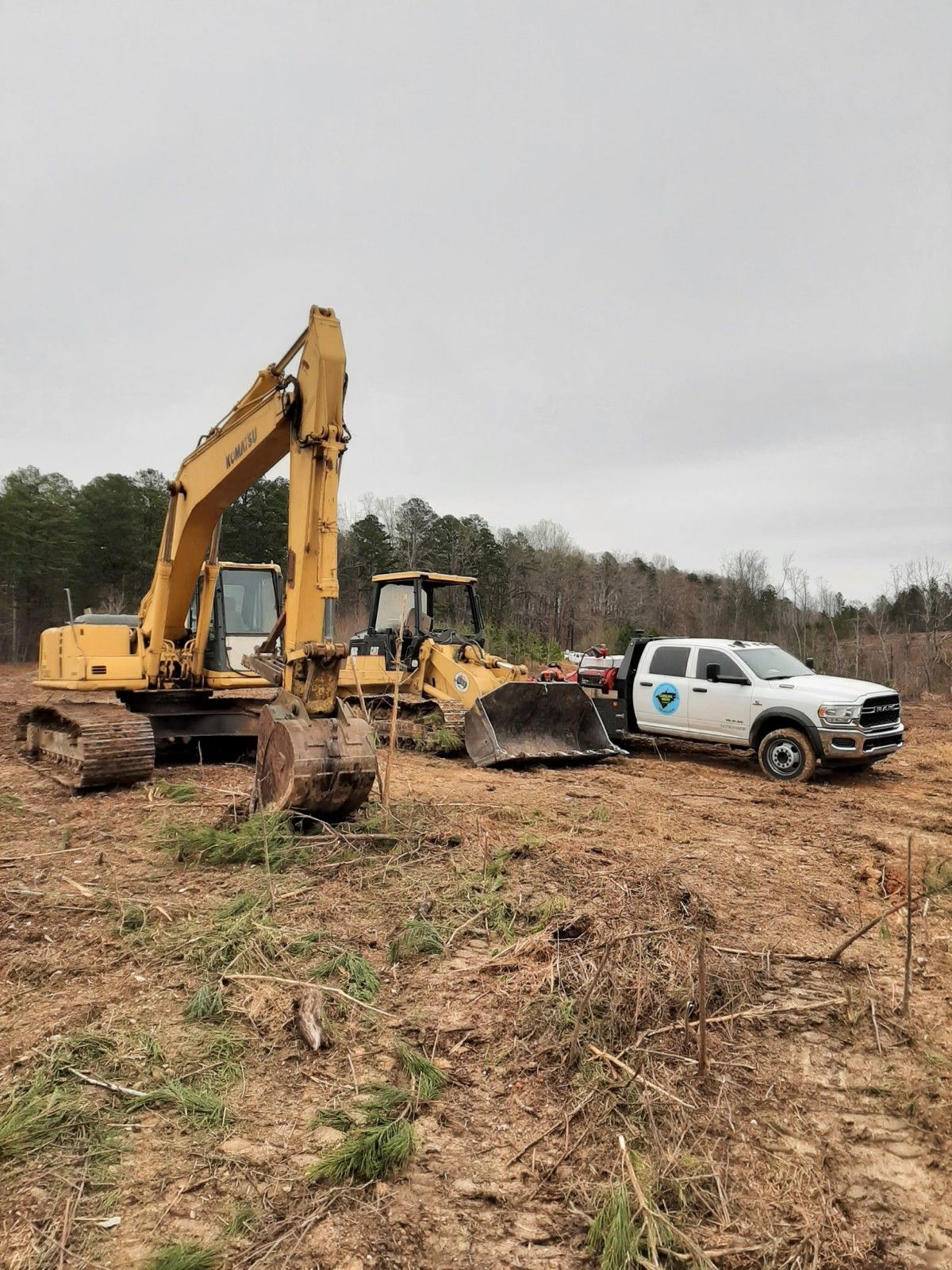 Yellow excavator and bulldozer clearing land, white truck with logo nearby. Cloudy day.