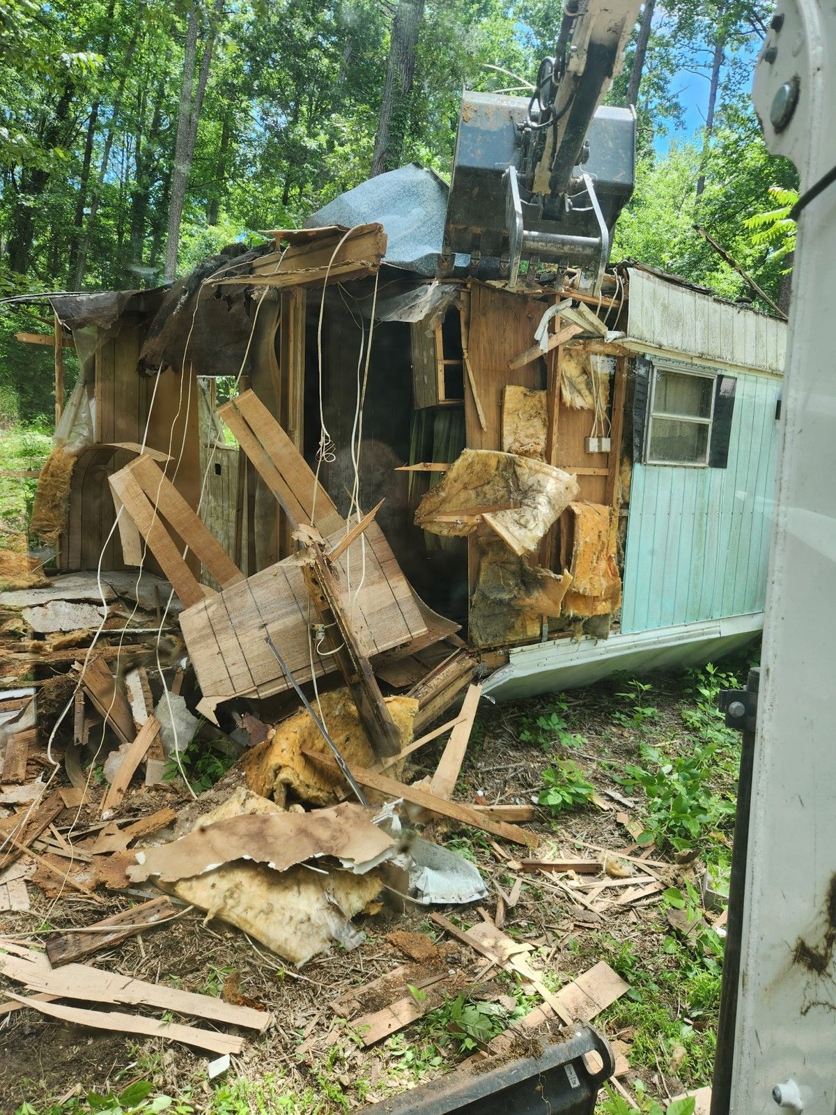 A dilapidated mobile home being demolished by an excavator in a wooded area. Debris is scattered.