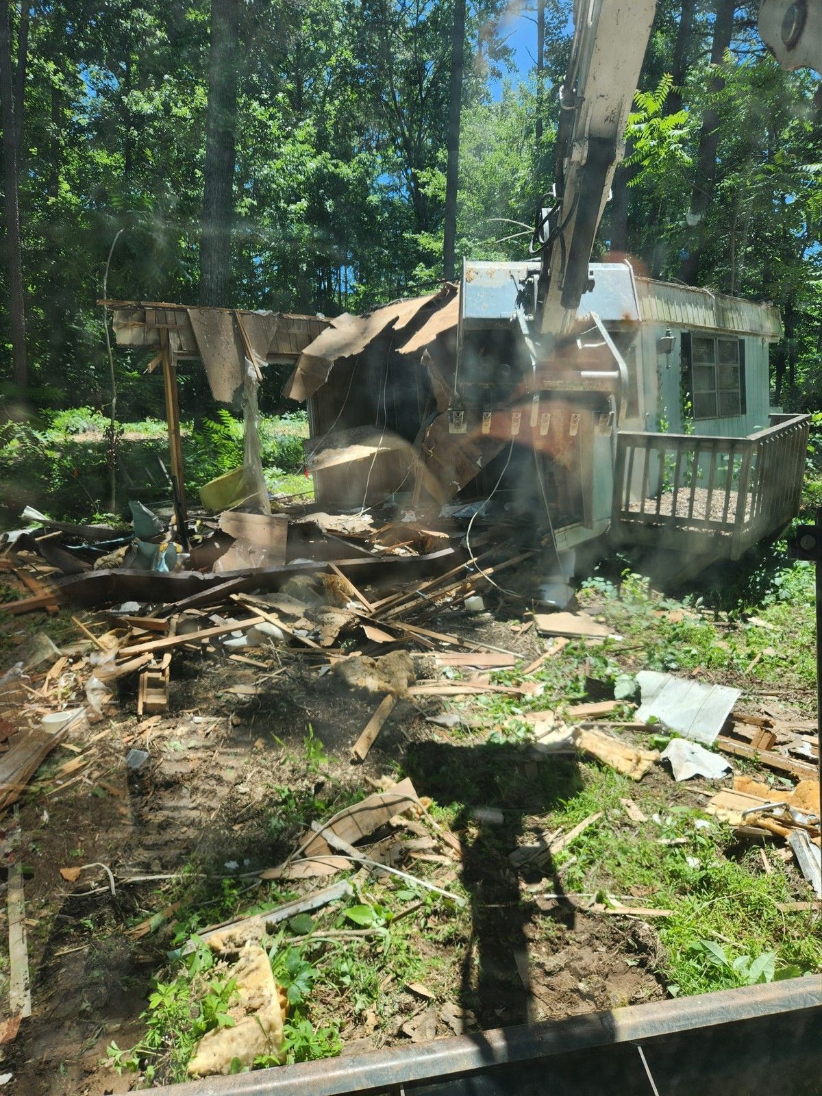 Demolition of small structure; excavator tears it down. Debris scattered on ground with trees in background.
