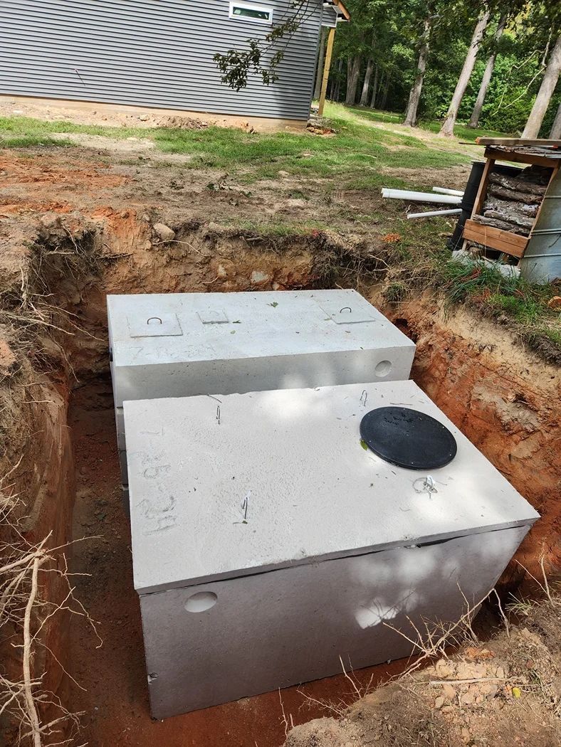 A concrete septic tank is set in a dirt excavation with a black circular lid, in a backyard setting.