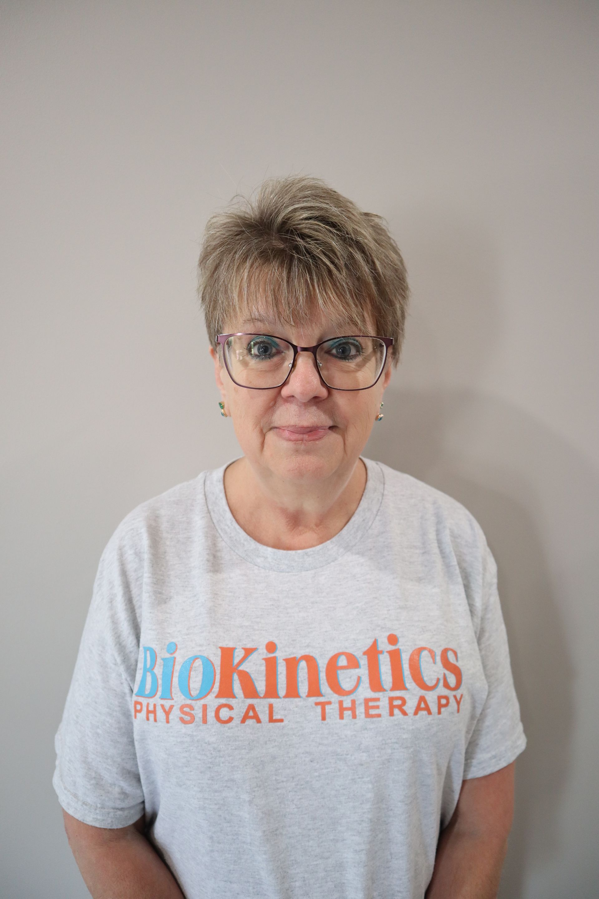 A woman wearing glasses and a biokinetics t-shirt is standing in front of a wall.