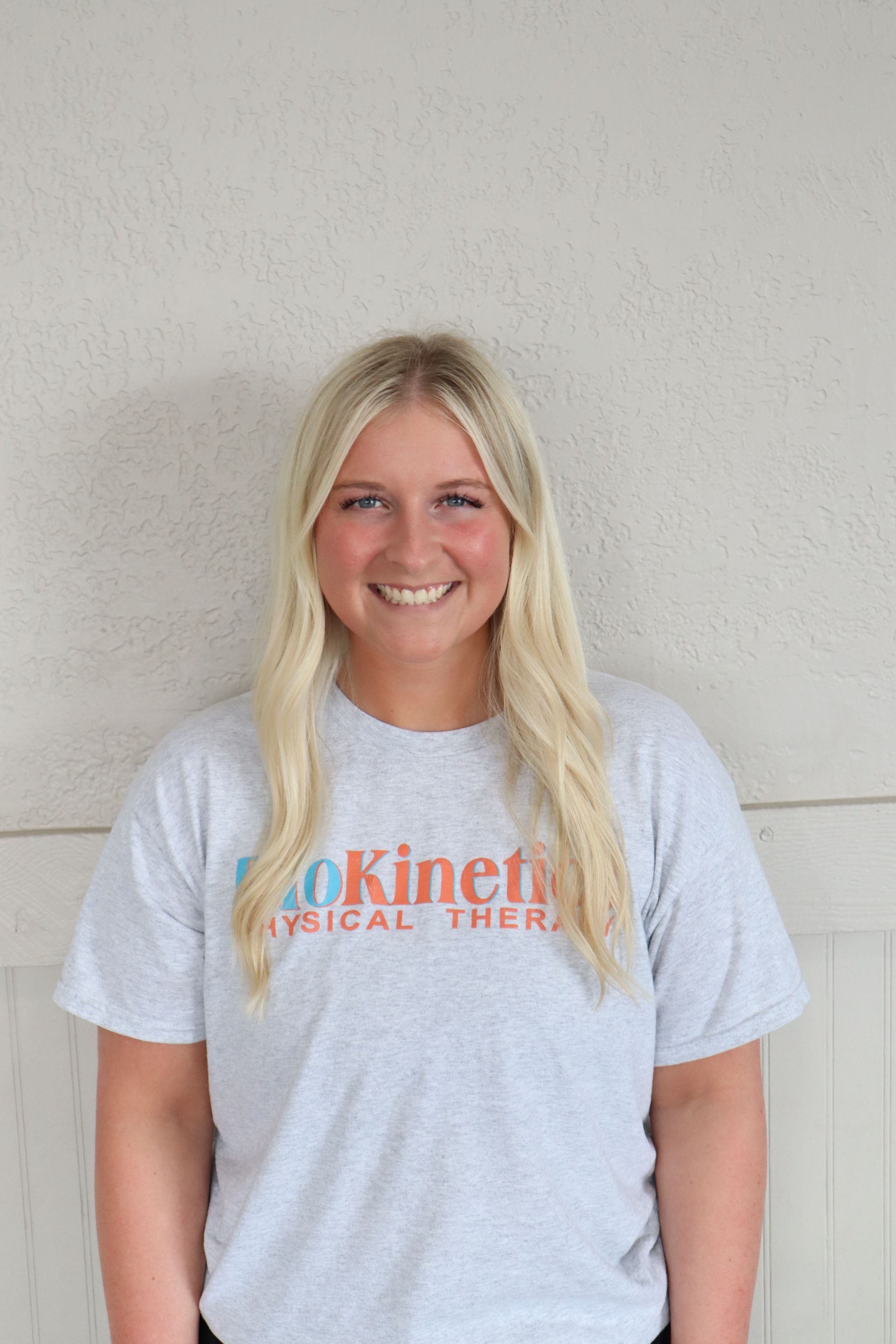 A woman wearing a gray t-shirt is smiling and standing in front of a wall.