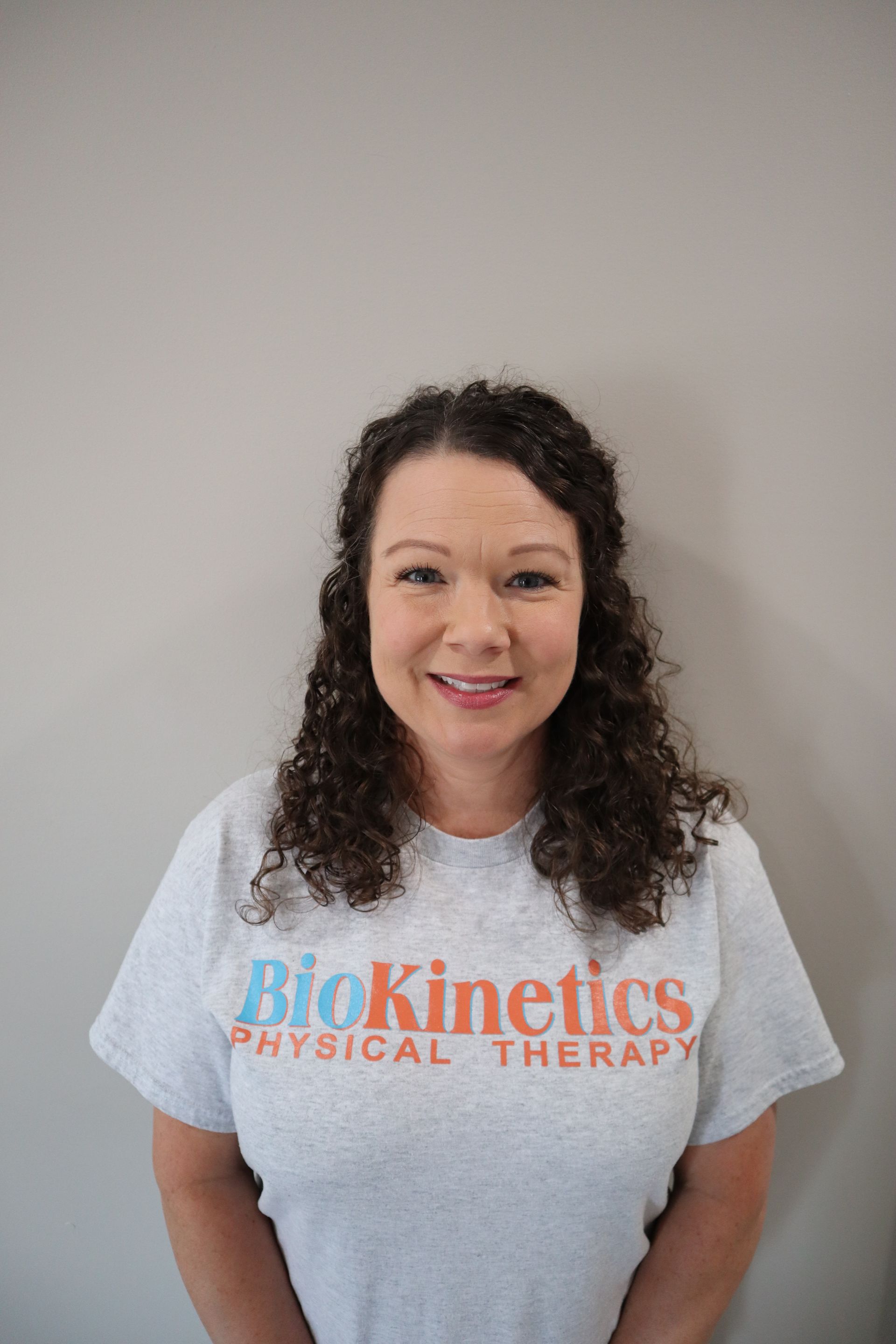 A woman is wearing a biokinetics physical therapy t-shirt and smiling.