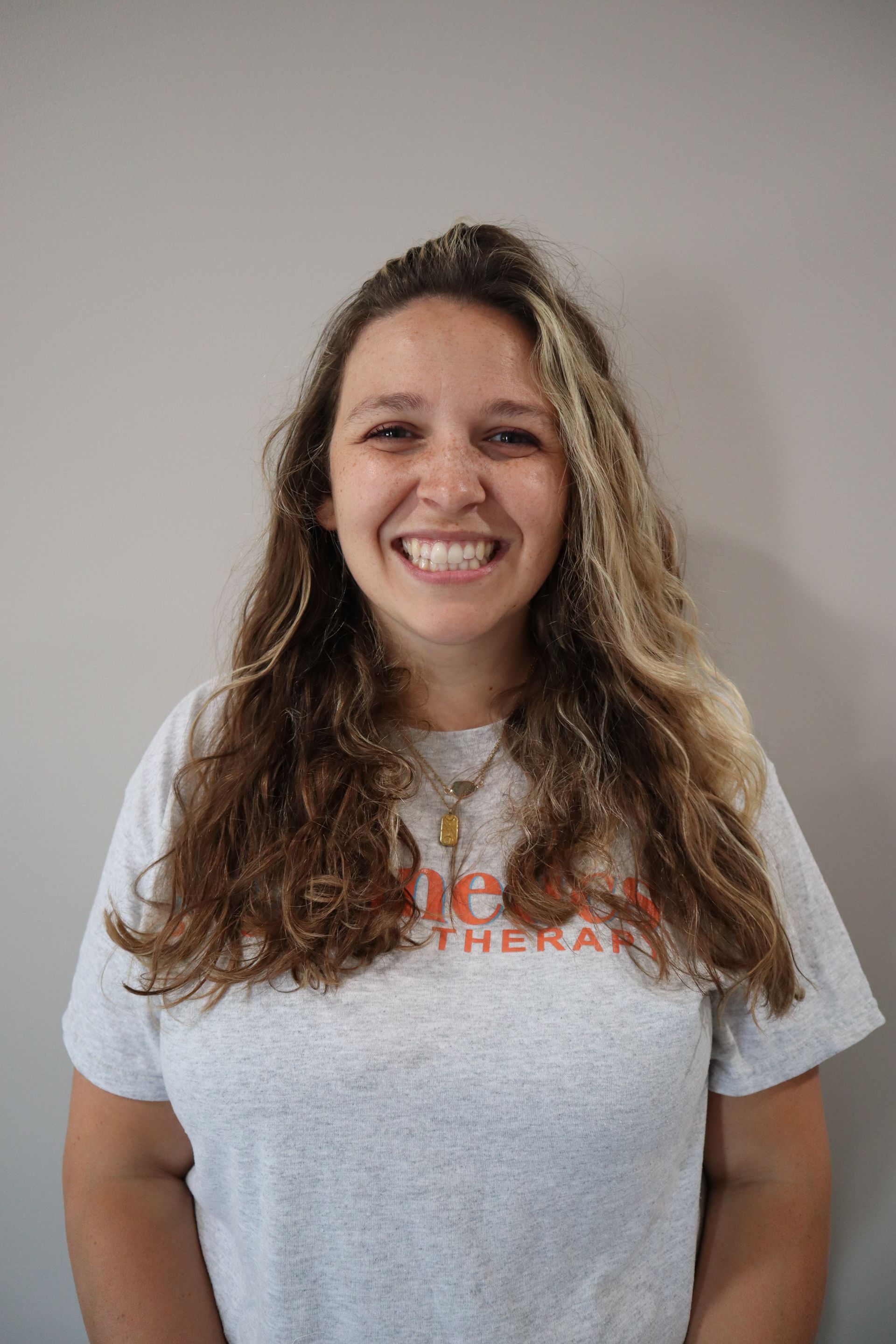 A woman with long hair is wearing a white t-shirt and smiling.