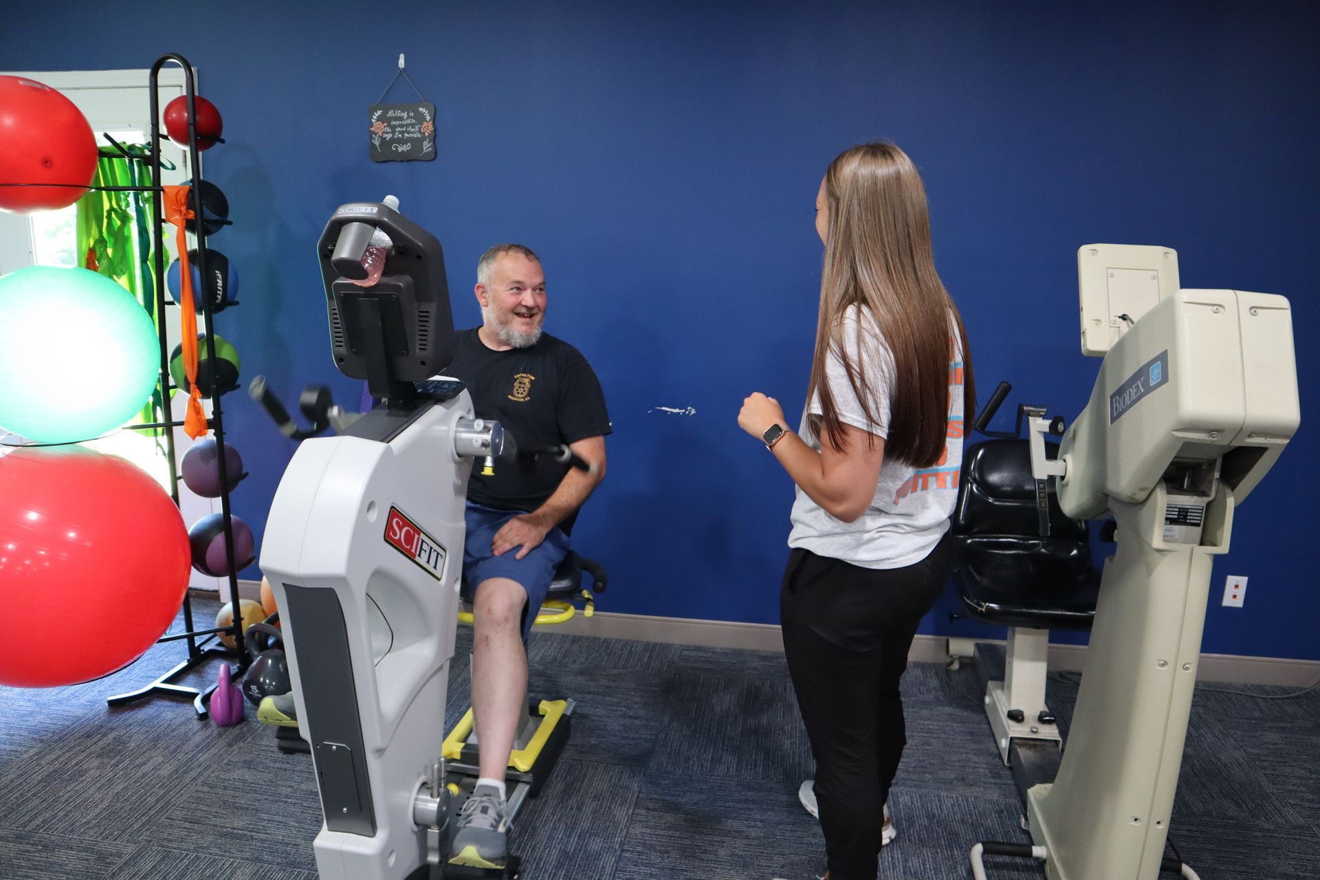 A man is riding an exercise bike in a gym while a woman stands next to him.