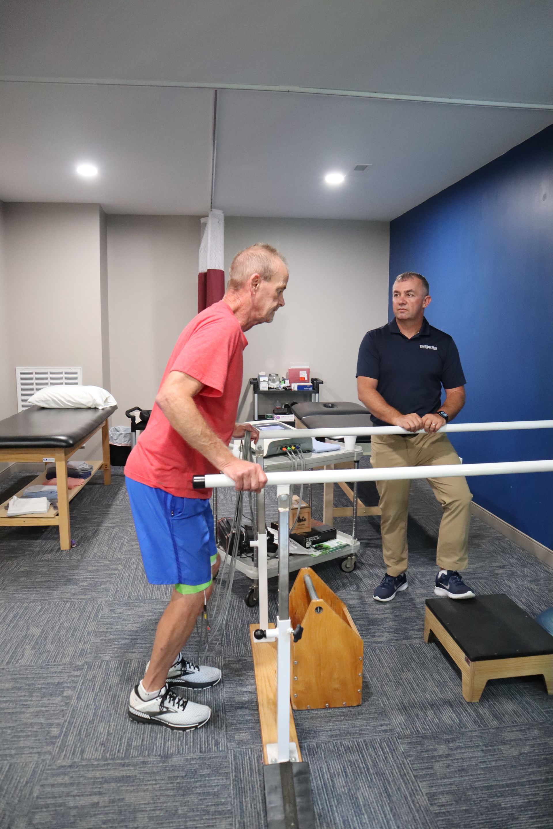 A man is walking on parallel bars in a therapy room.