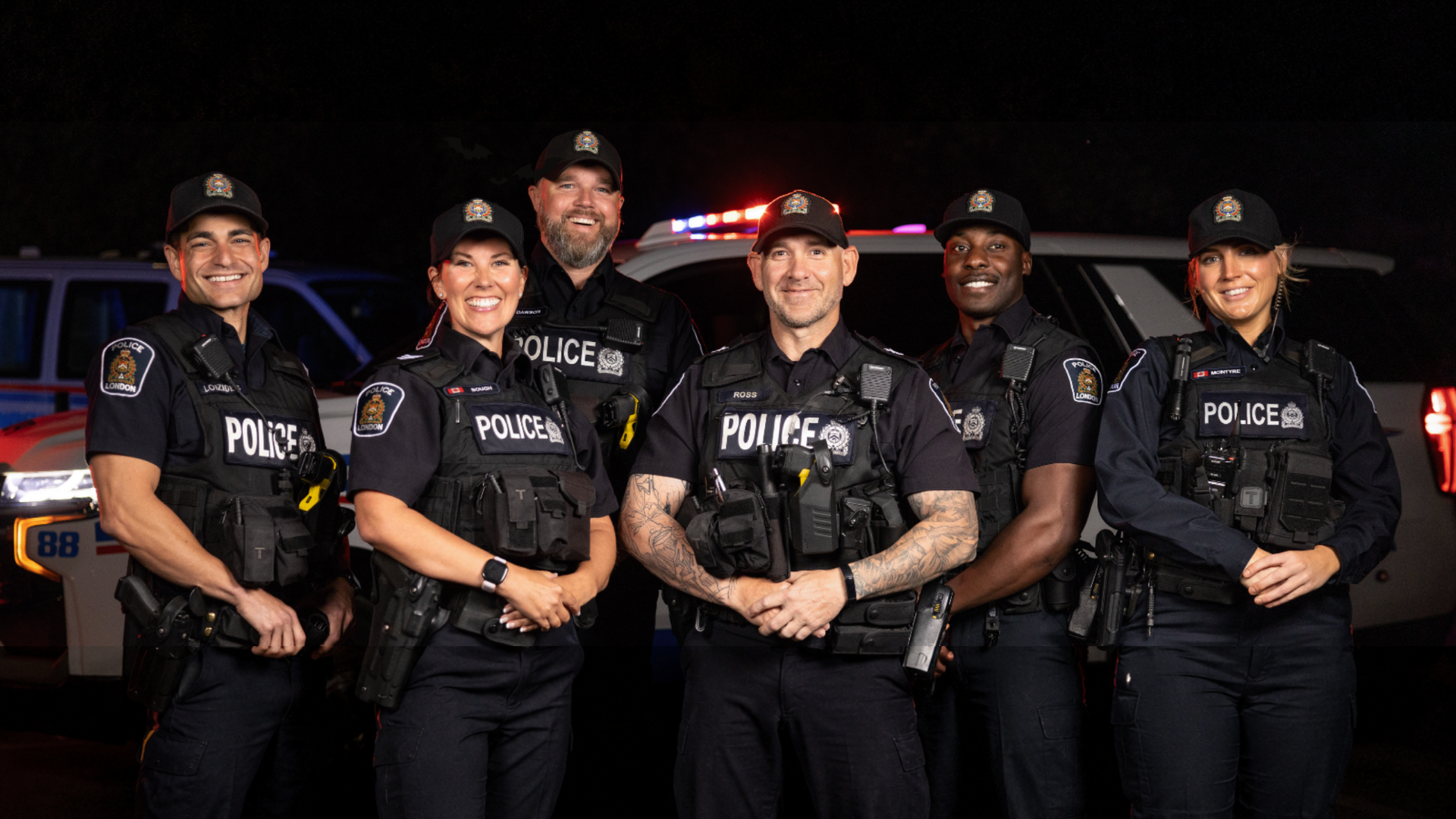 Six police officers in black uniforms standing together at night, smiling in front of patrol vehicles.