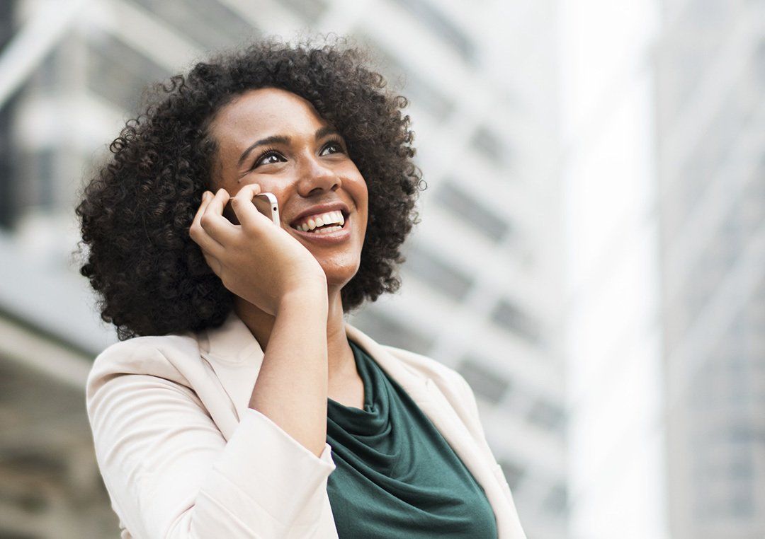 Woman smiles while talking on a phone outdoors, near a tall building.
