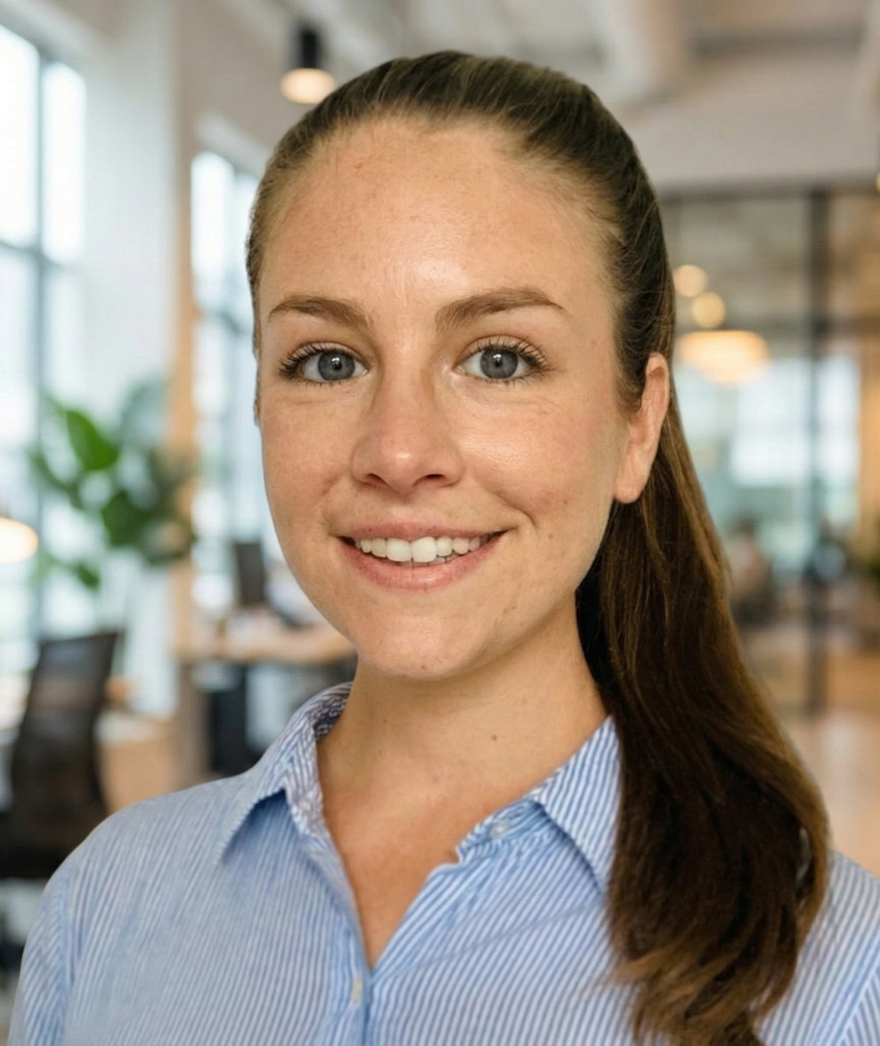 Woman in blue striped shirt smiles in an office setting, ponytail, blurred background.