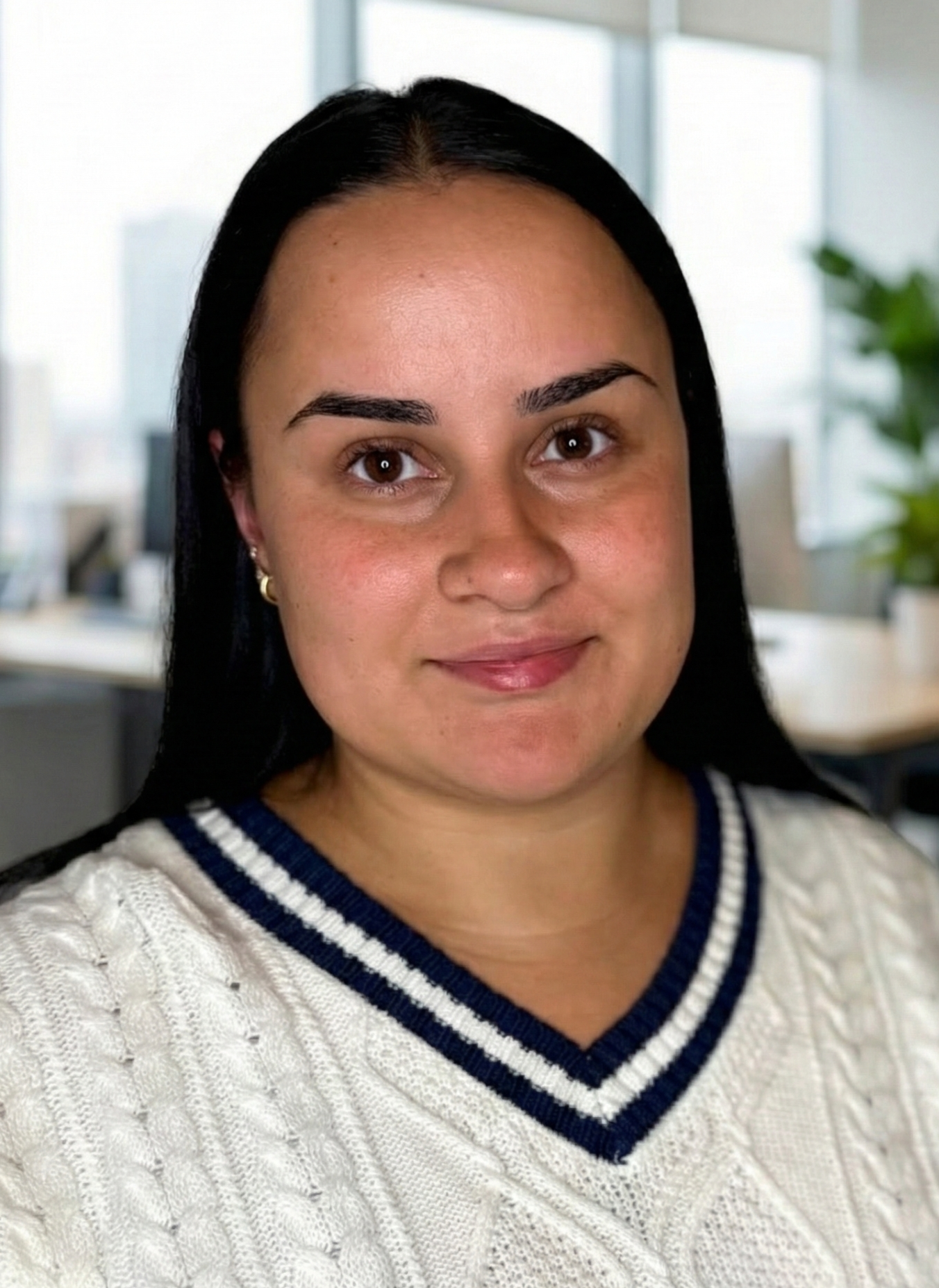 Woman with dark hair smiles, wearing a cream sweater with a blue stripe, in an office setting.