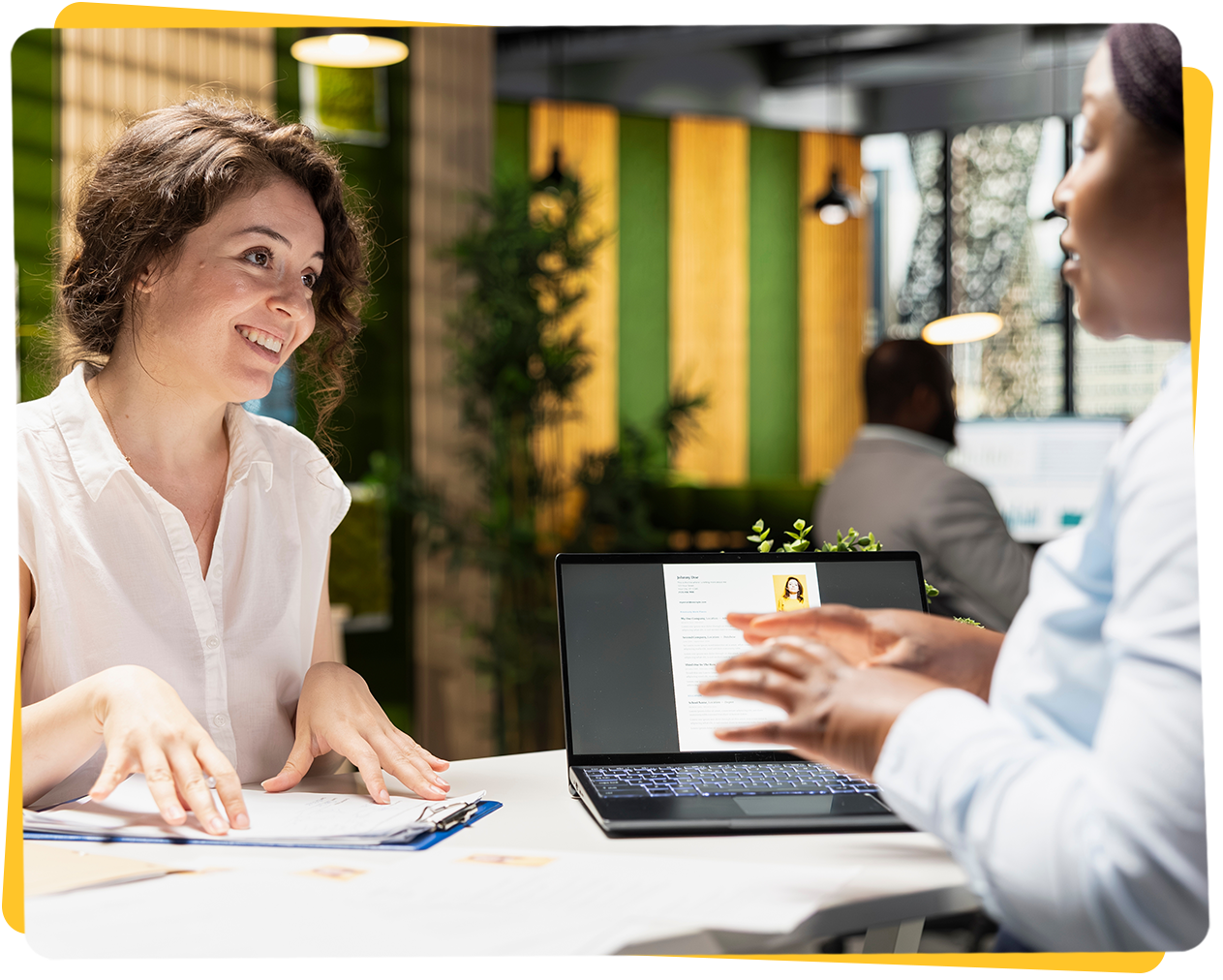 Woman smiling and gesturing while reviewing documents with another person at a table, laptop in between.