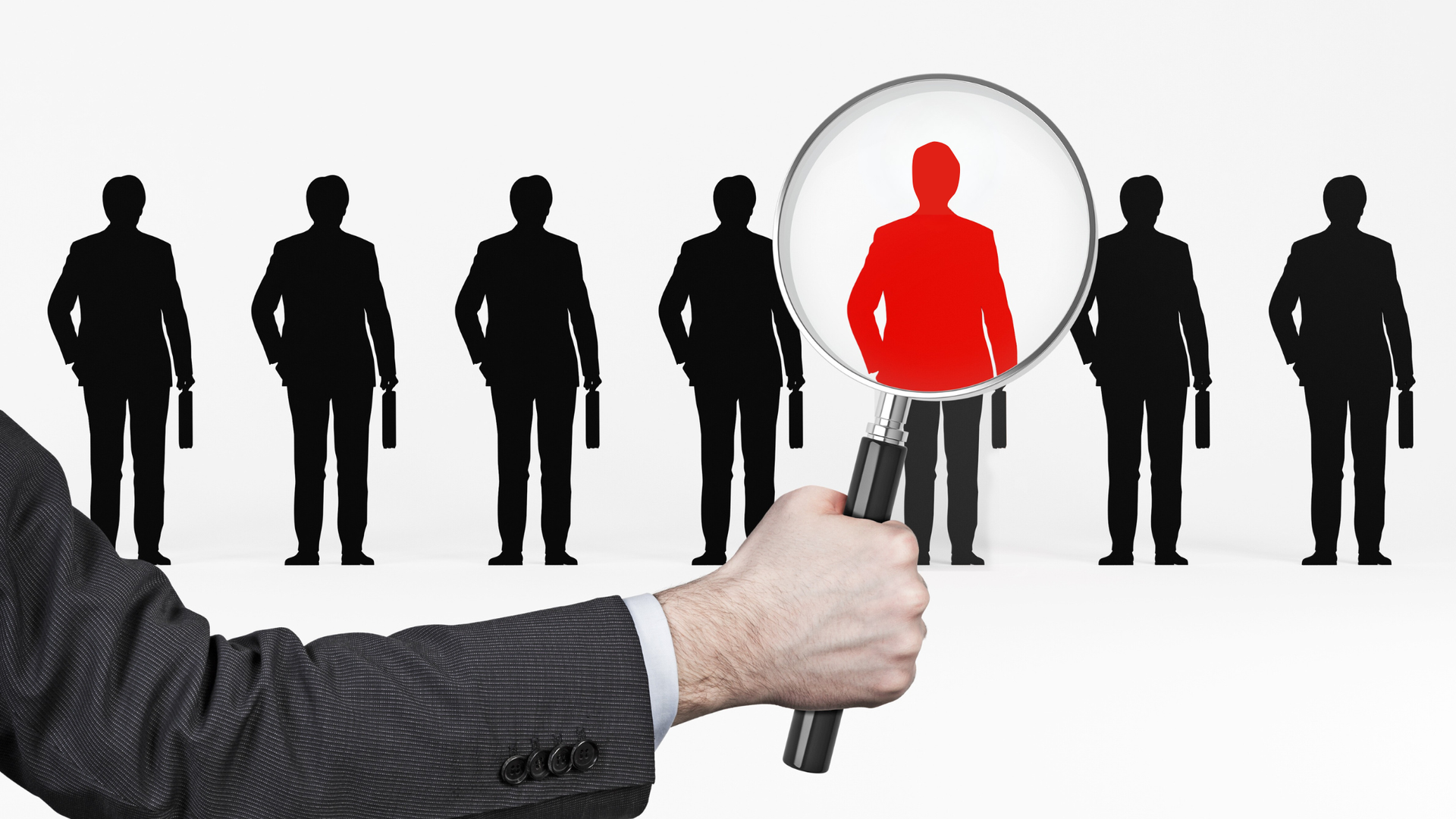 Hand holding magnifying glass highlighting a red silhouette of a person among black silhouettes.