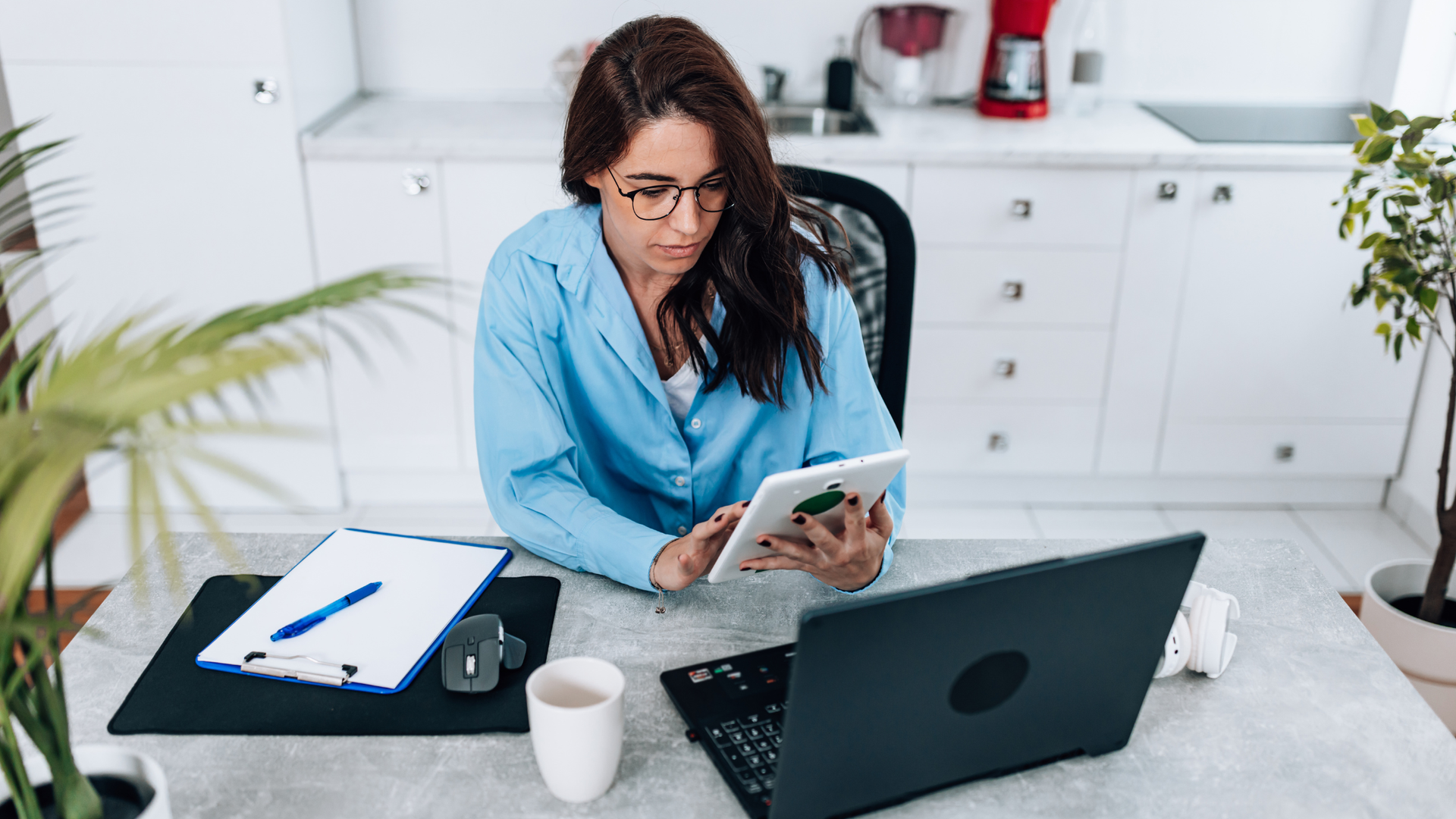 Woman in a blue shirt taking notes while reading a tablet at a desk with a laptop in a bright office