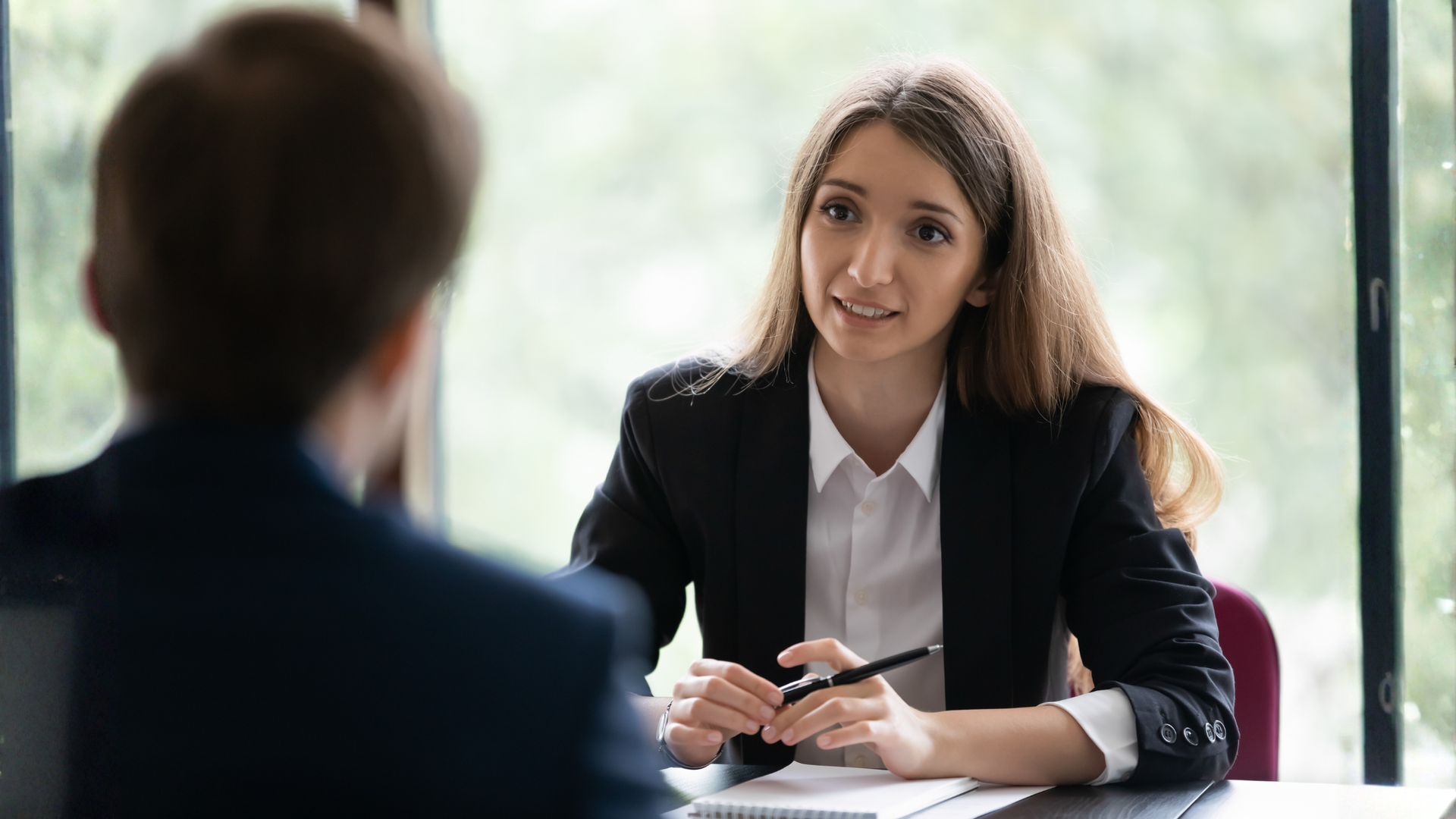 Woman in suit speaking to person at a table, natural light, holding pen, serious expression.