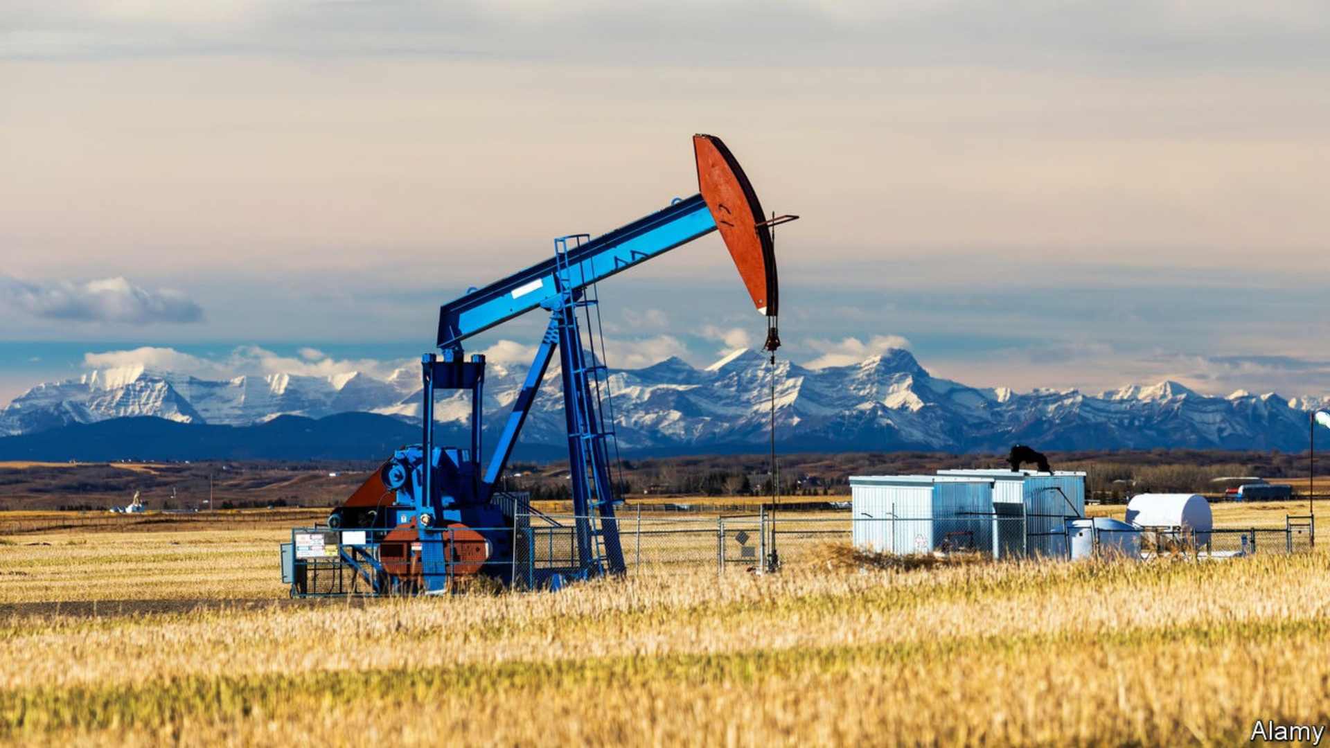 A blue oil pump jack operates in a dry, golden field with snow-capped mountains in the distant background.