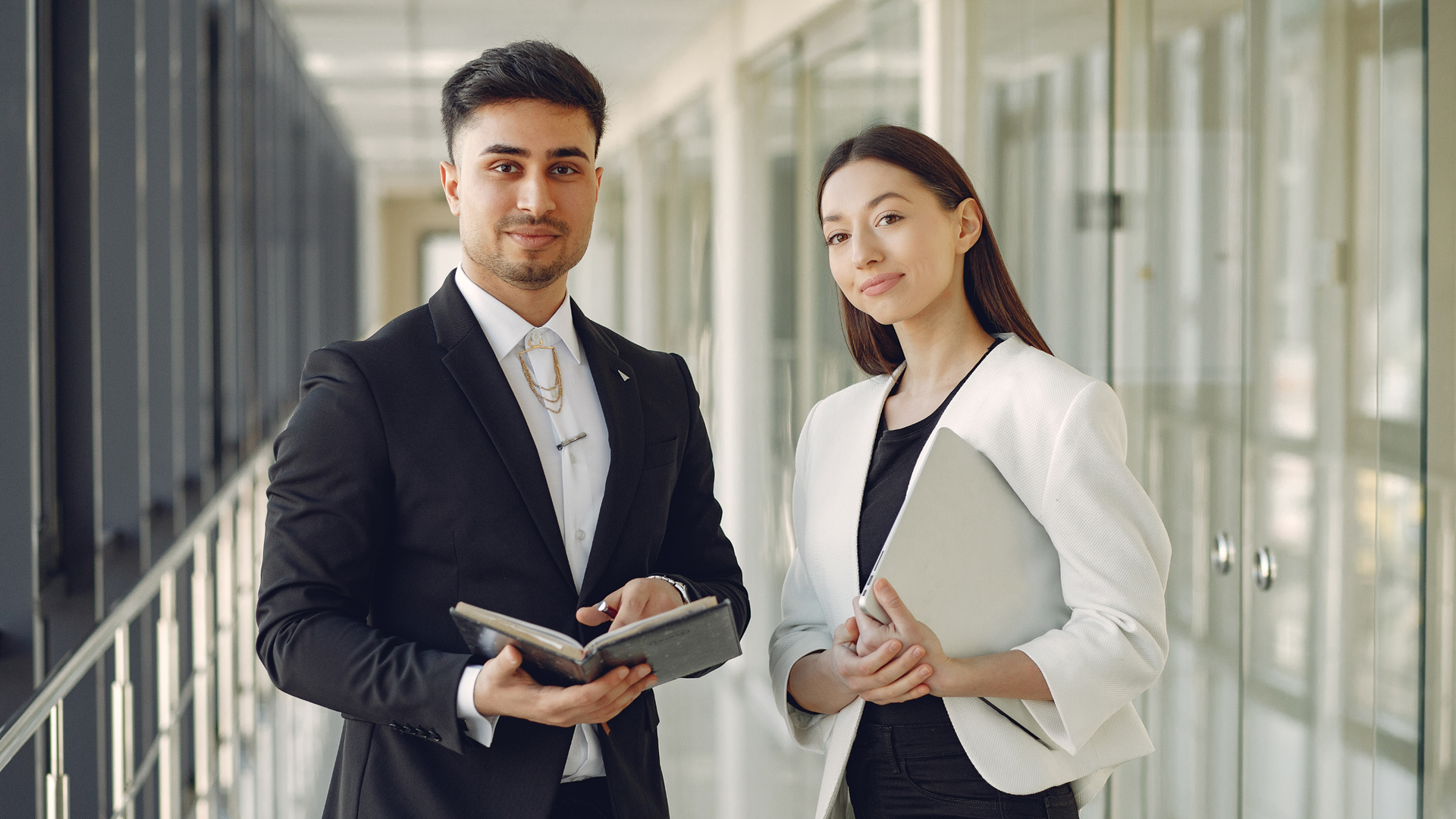 Man and woman in business attire, looking toward camera in office hallway, man holding notebook, woman holding laptop.