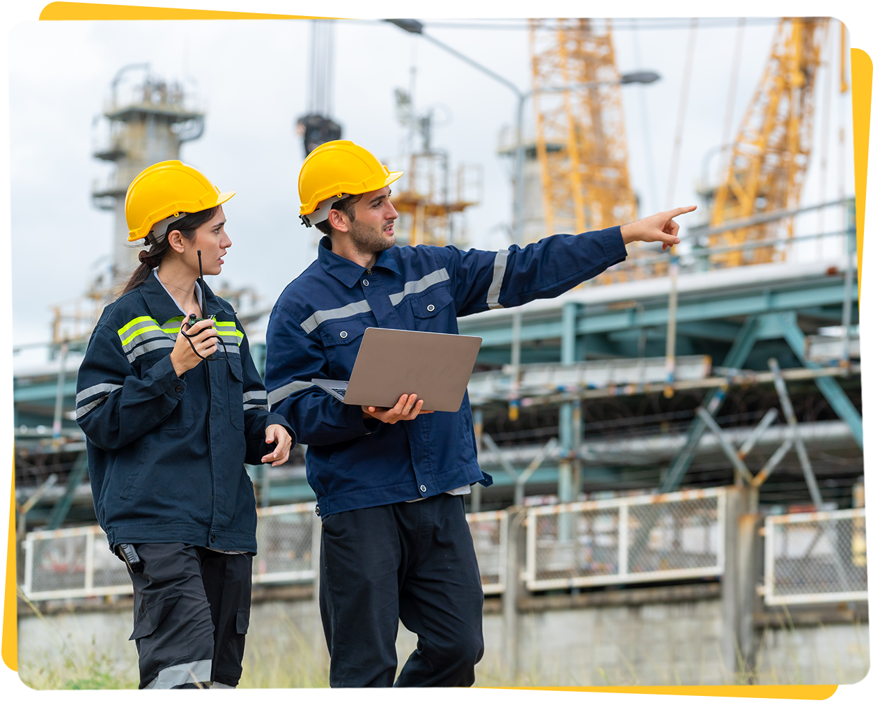 Two people in hard hats and workwear at an industrial site; man points, woman looks on.