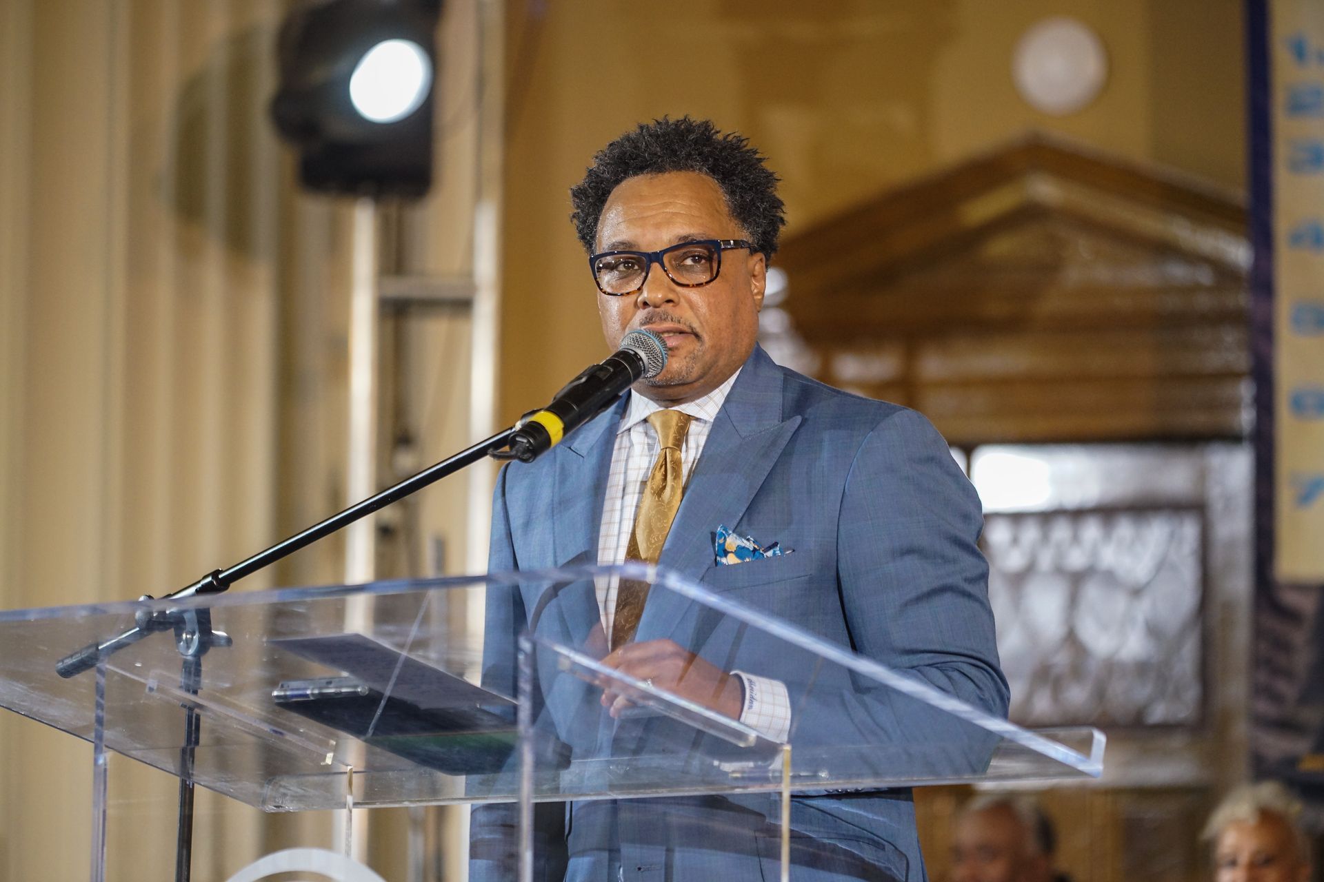 A man in a suit and tie is standing at a podium speaking into a microphone.