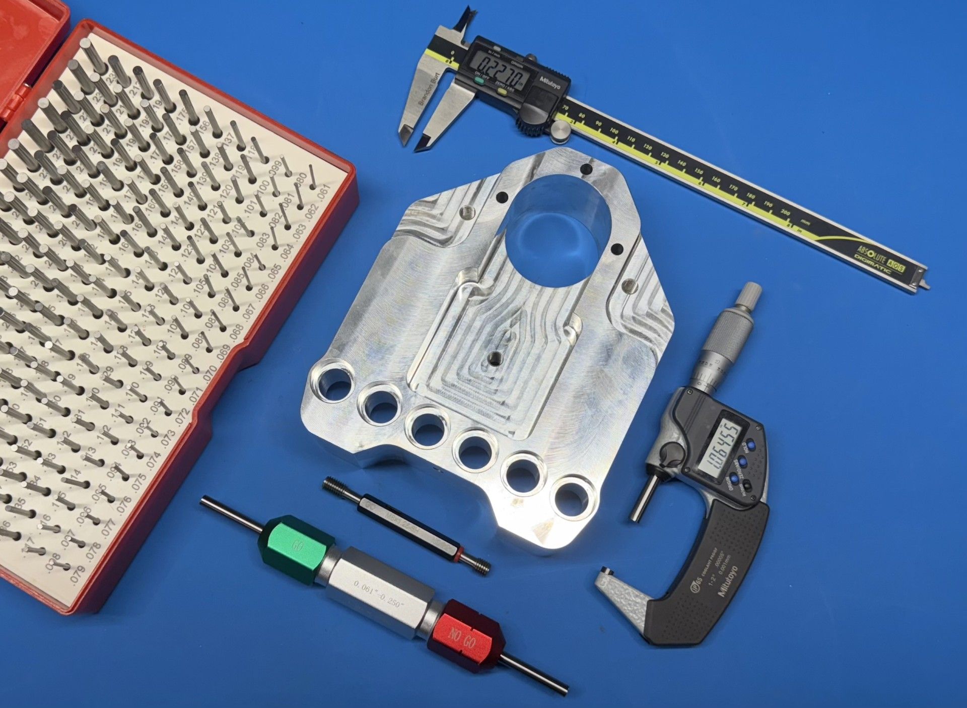 A metal part with precision measuring tools on a blue surface: calipers, micrometer, and pin gauges.