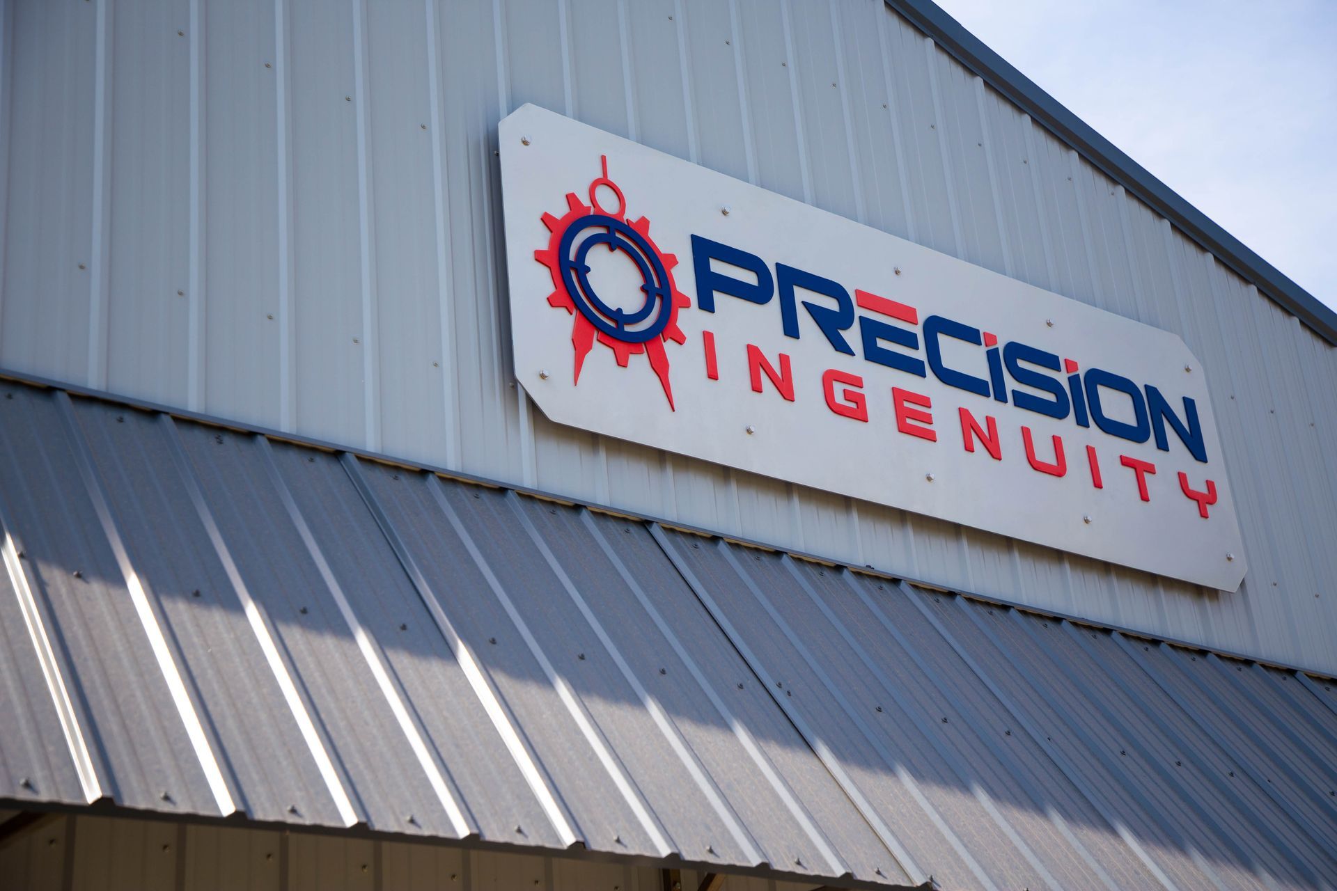 Sign for Precision Ingenuity on a white building. The logo features a blue gear, red rays, and blue text.