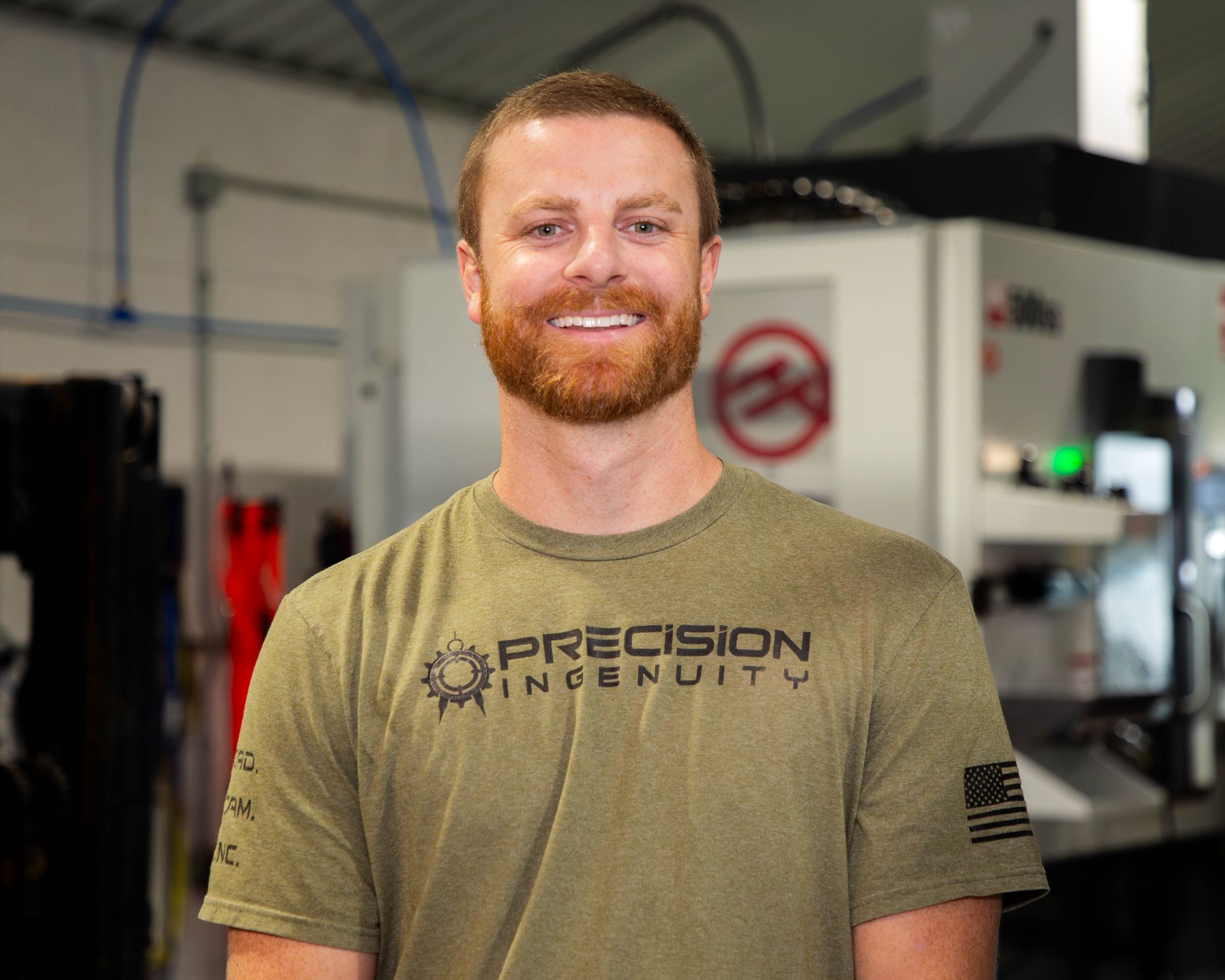Man in olive green shirt smiles in a workshop. Precision Ingenuity logo and American flag visible.