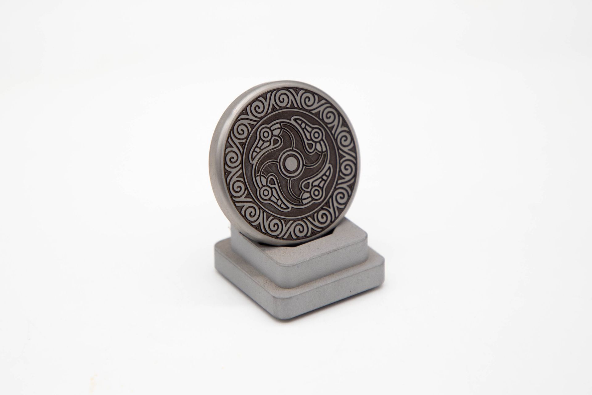 Metal coin with Celtic design on a gray pedestal, set against a white background.