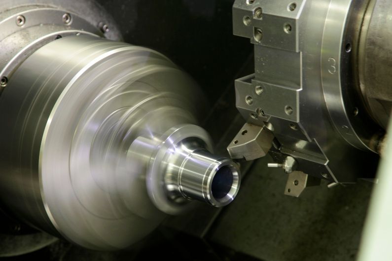 A metal lathe cutting a cylindrical metal part. The part spins rapidly while a cutting tool shapes it.