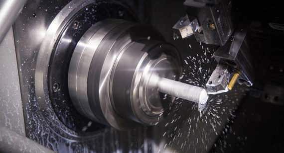 A CNC machine cutting a metal rod; metallic silver with cutting fluid spray in a workshop.