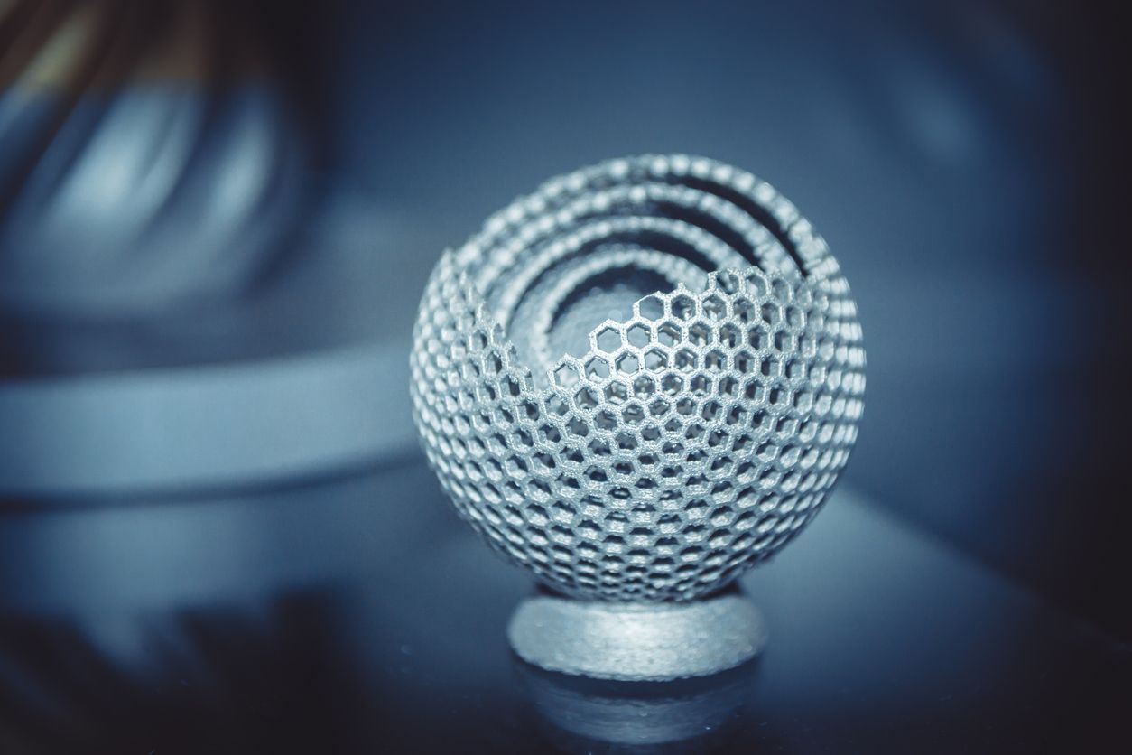 Honeycomb-patterned metal sphere sculpture, nested rings, resting on a reflective surface.