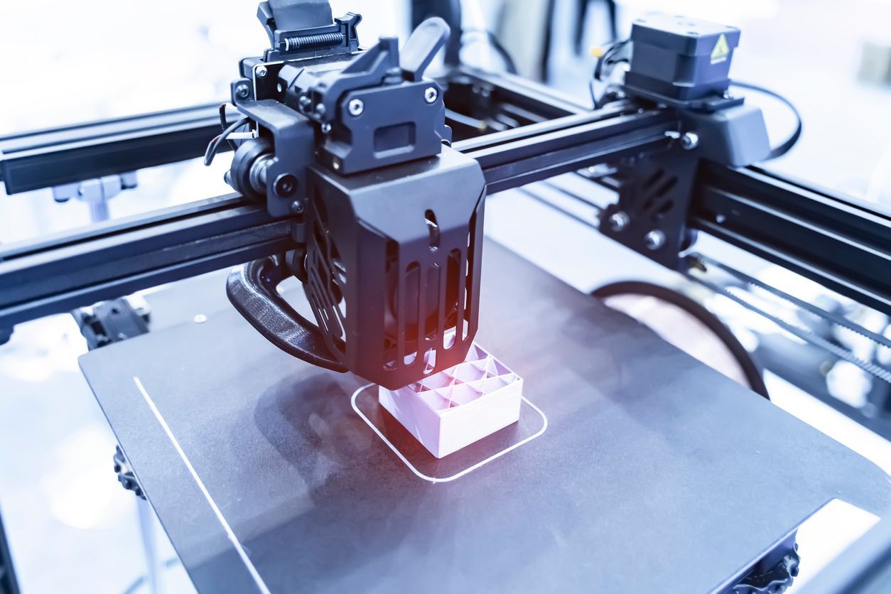 3D printer creating a pink, rectangular object on a gray base. The machine is black with visible mechanics.