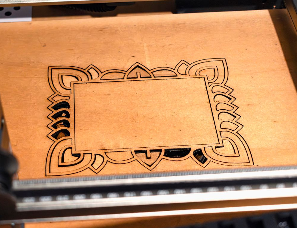 Laser cutter etching an ornate floral frame design onto a wooden board.
