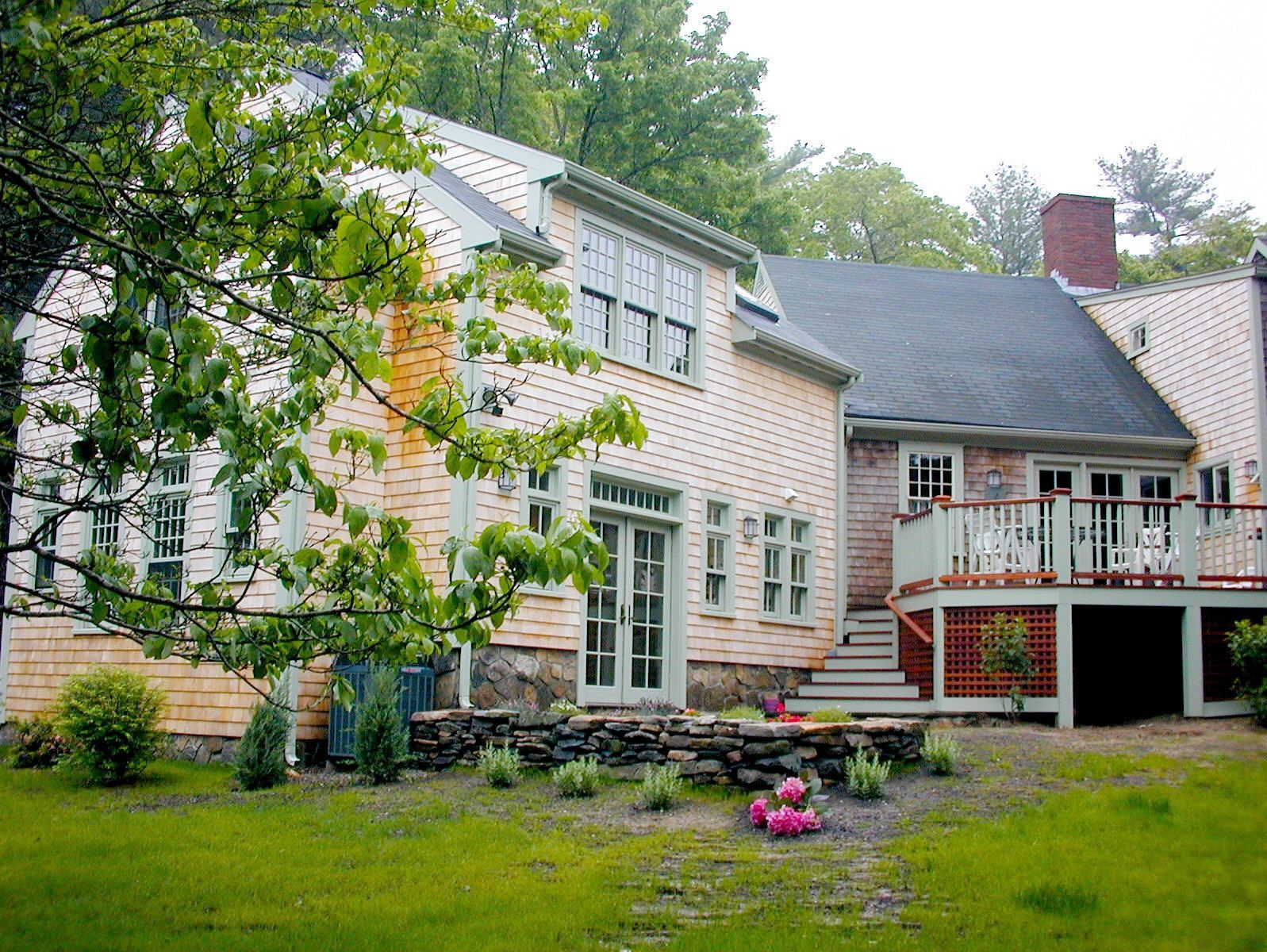 Beige shingled house with light green trim and a wooden deck. Lush greenery surrounds.