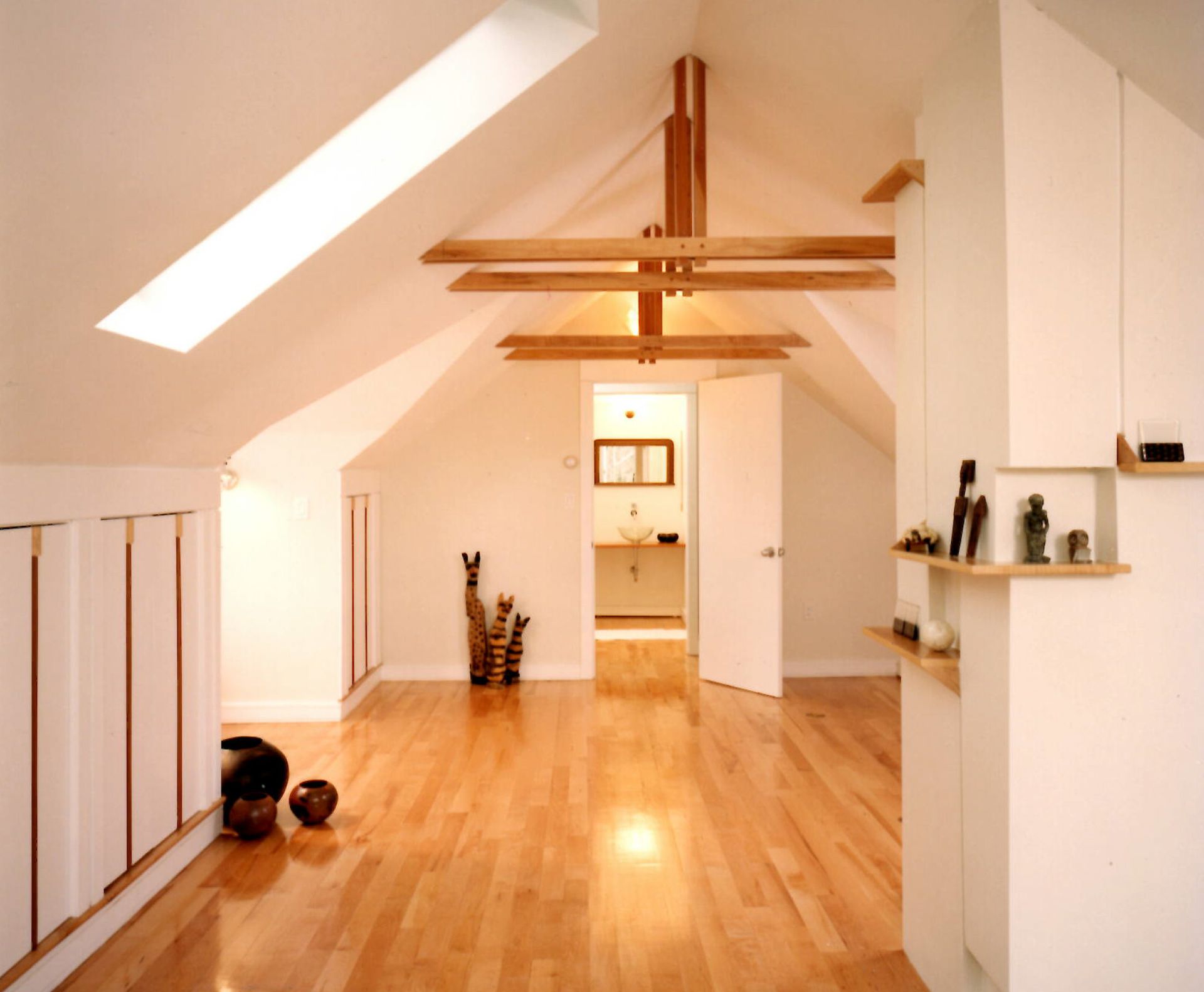 Bright, airy loft space with wooden floors, white walls, and a skylight. Features hanging wooden beams and open doorway.