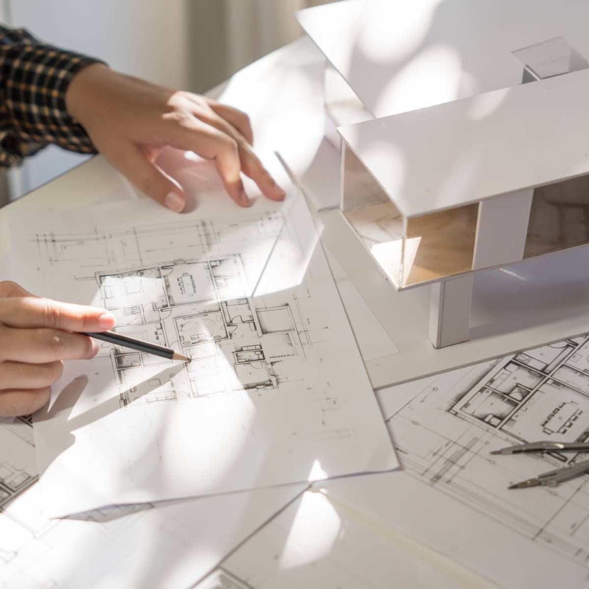 Architect's hand pointing at blueprint, next to a model house and drafting tools.