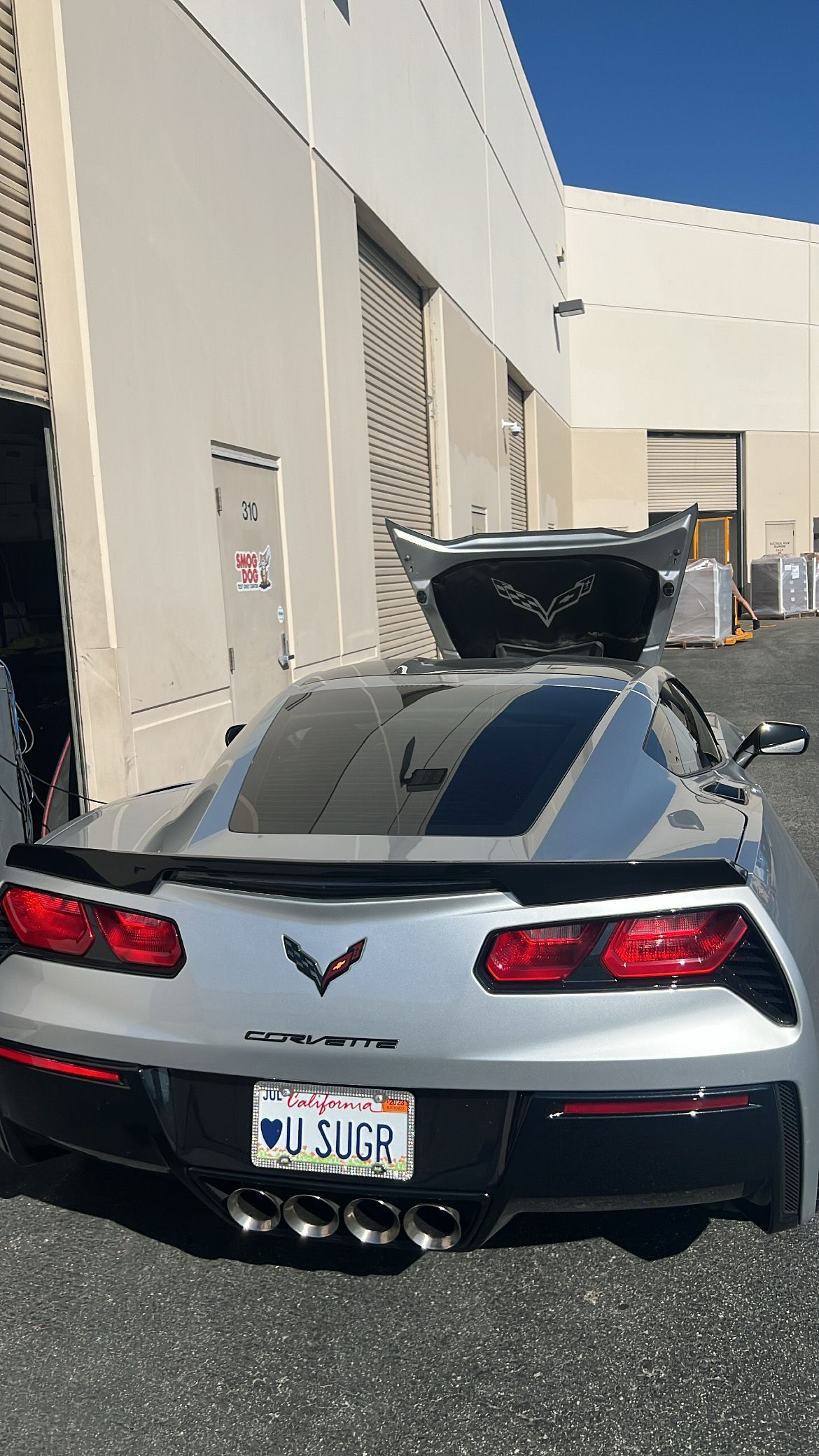 A silver corvette with the hood up is parked in front of a building.