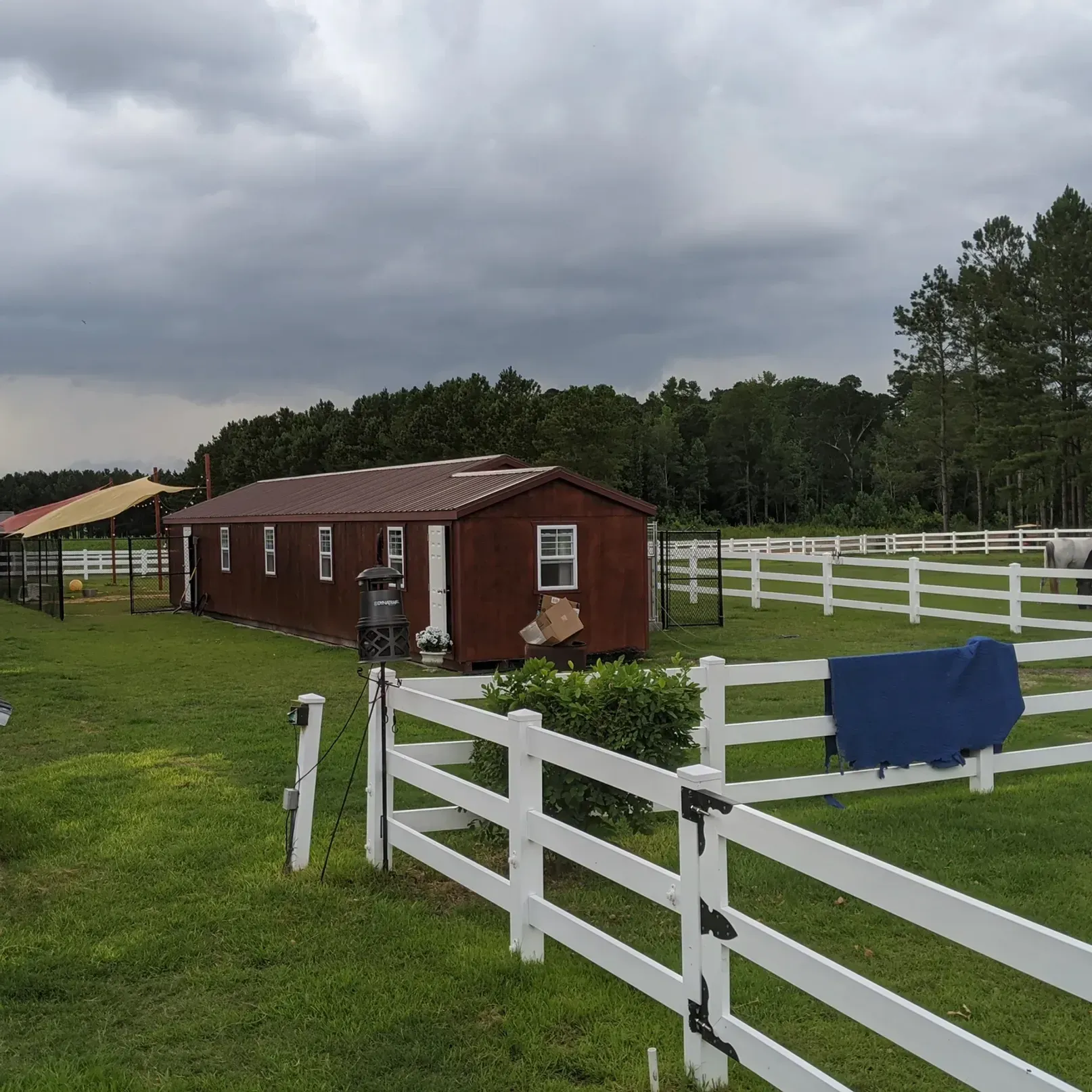 Red building with white fences and fields, against a treeline and cloudy sky.