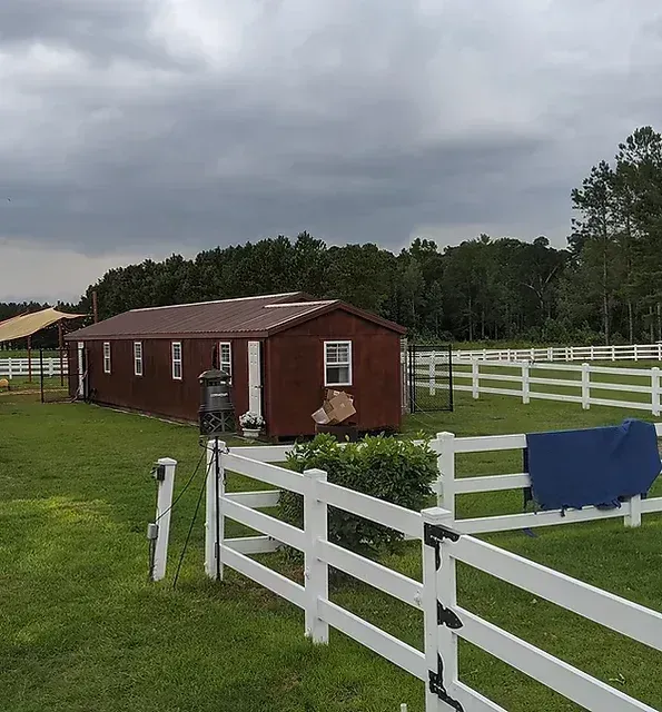 Red barn with white fences on a grassy field, under an overcast sky.